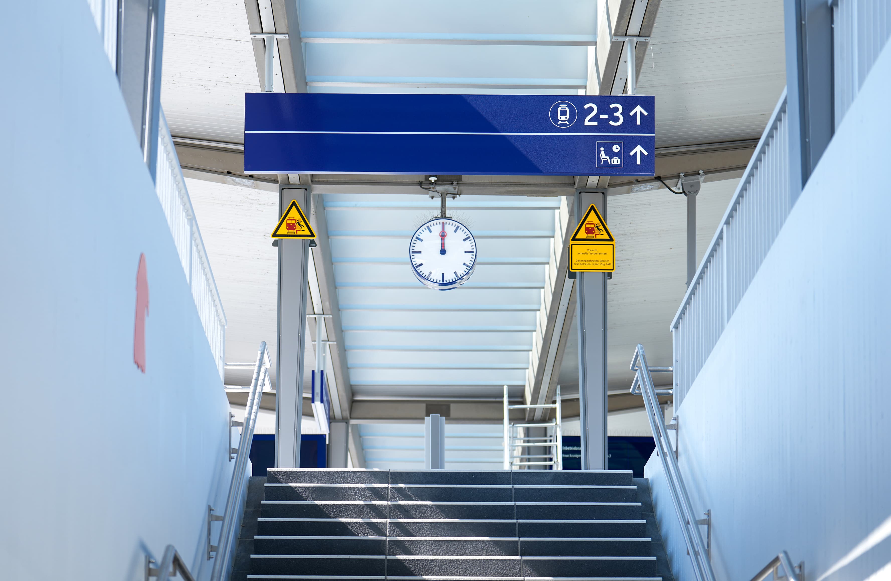 Eine Treppe, eine Uhr und blauer Wegweiser an einem Bahnhof.