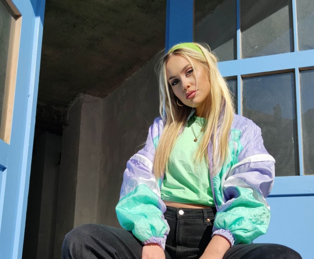 Katharina Engel sitzt auf einem roten Stuhl vor blauen Türen und trägt eine bunte Jacke, grünes Shirt, schwarze Hose und schwarze Sneakers