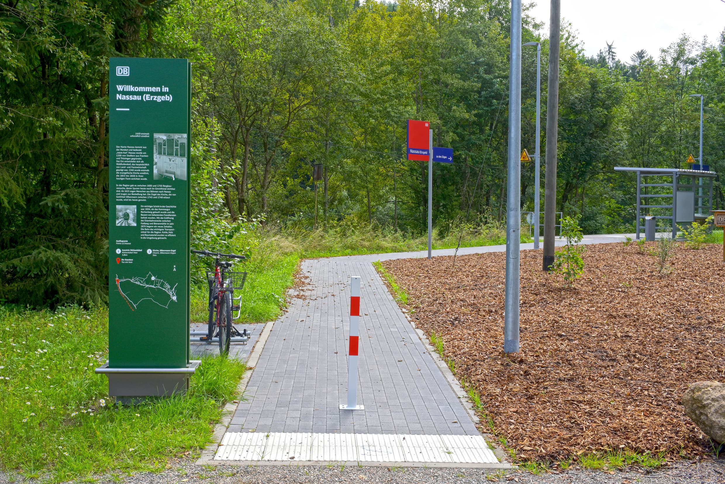 Zu sehen ist die Zuwegung zum Bahnhof, links neben des Weges steht eine große, grüne Stele mit Informationen zur Kommune Nassau und einfache Fahrradbügeln. Rechts neben dem Weg wurde eine Grünfläche angelegt auf der die ersten kleineren Pflanzen zu erkennen sind.