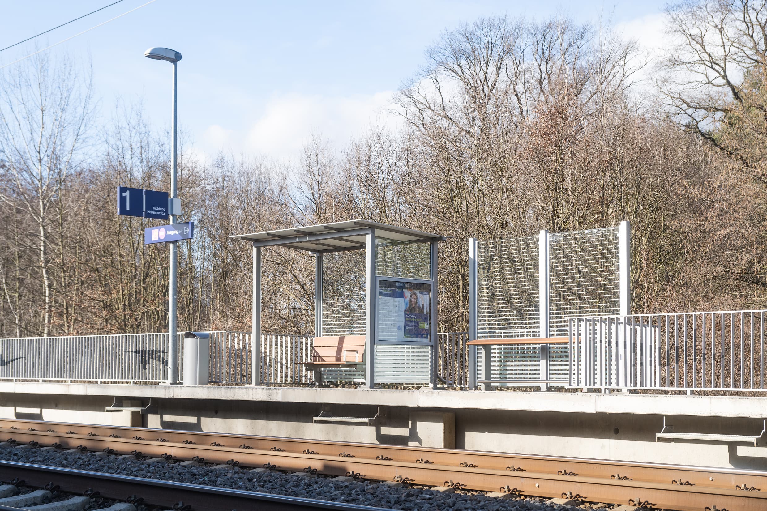 Blick auf den Bahnsteig und das volltransparente Wetterschutzhaus mit einer neuen Holzbank. Rechts daneben wurde ein zusätzlicher Anlehner installiert. Außerdem erkennt man zwei Vogelsilhouetten, die malerisch auf das Geländer angebracht wurden.