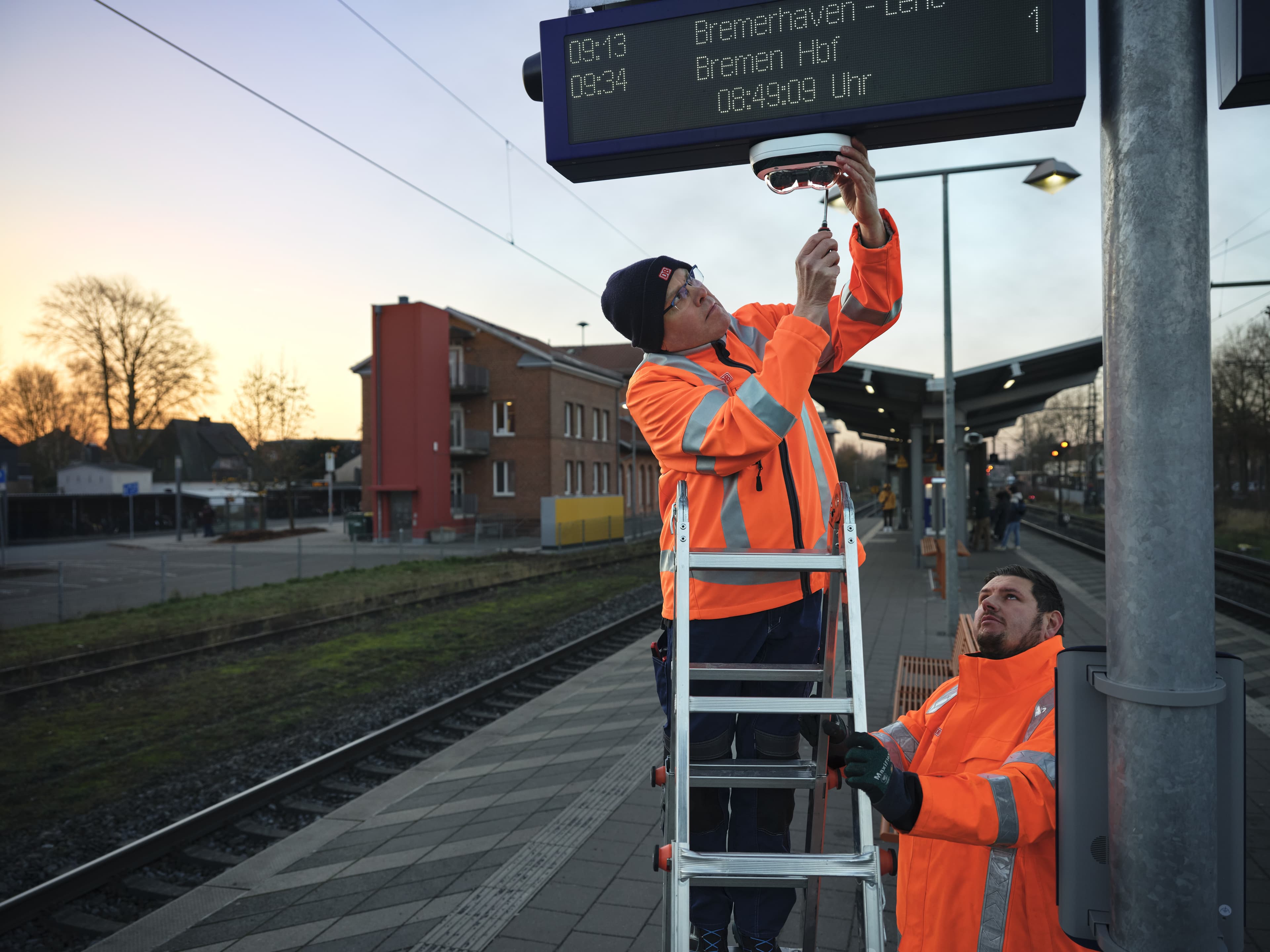 Ein Team aus zwei Männern montiert gemeinsam eine neue Videoüberwachungsanlage an einem Bahnsteig.