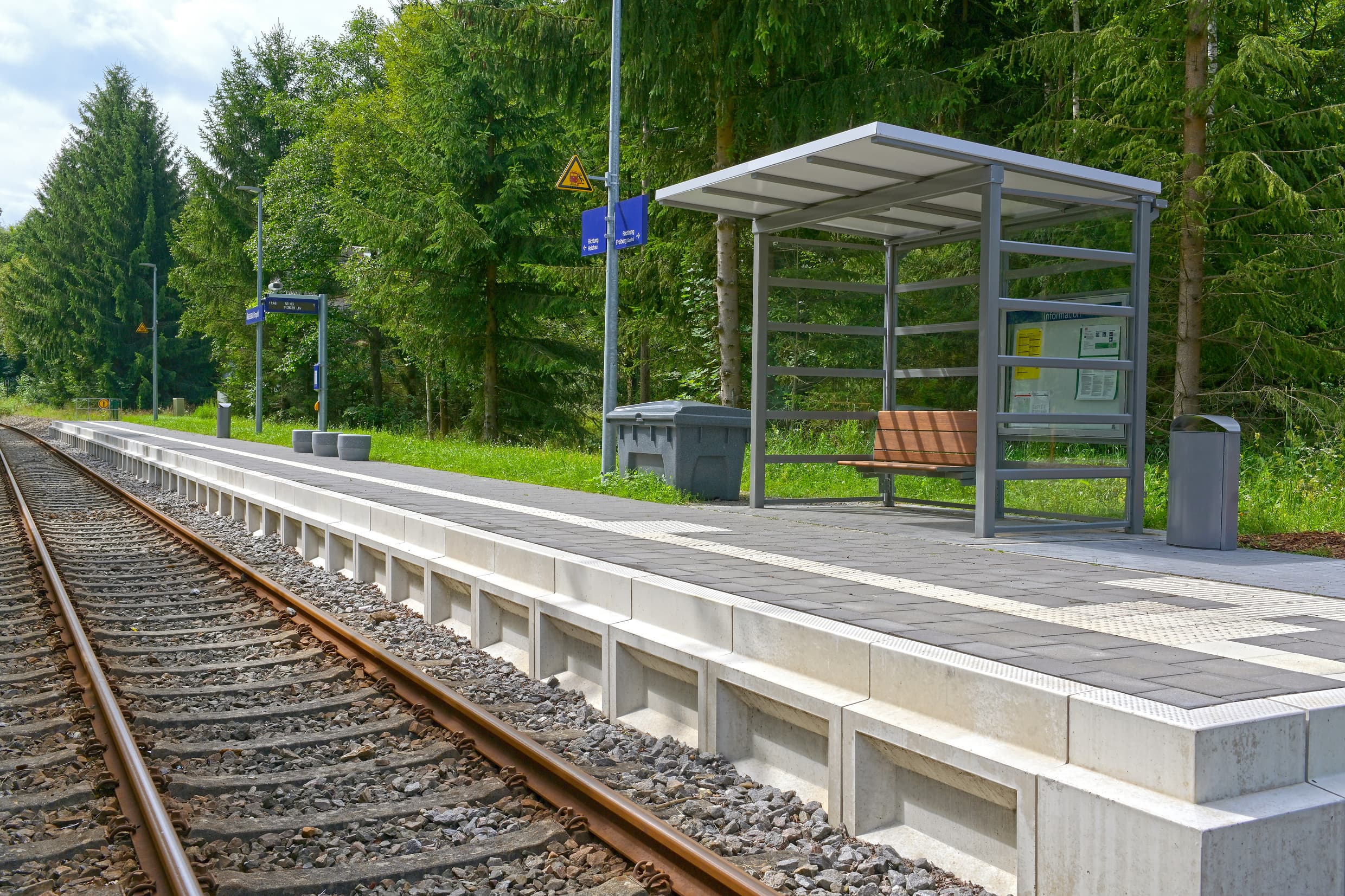 Zu sehen ist ein neu gebauter Bahnsteig mit neuer, moderner Ausstattung wie drei Betonsitzhockern, einer digitalen Reisendeninformation, ein Streugutbehälter links neben dem volltransparenten Wetterschutzhaus mit einer Vitrine und einer Holzsitzbank.