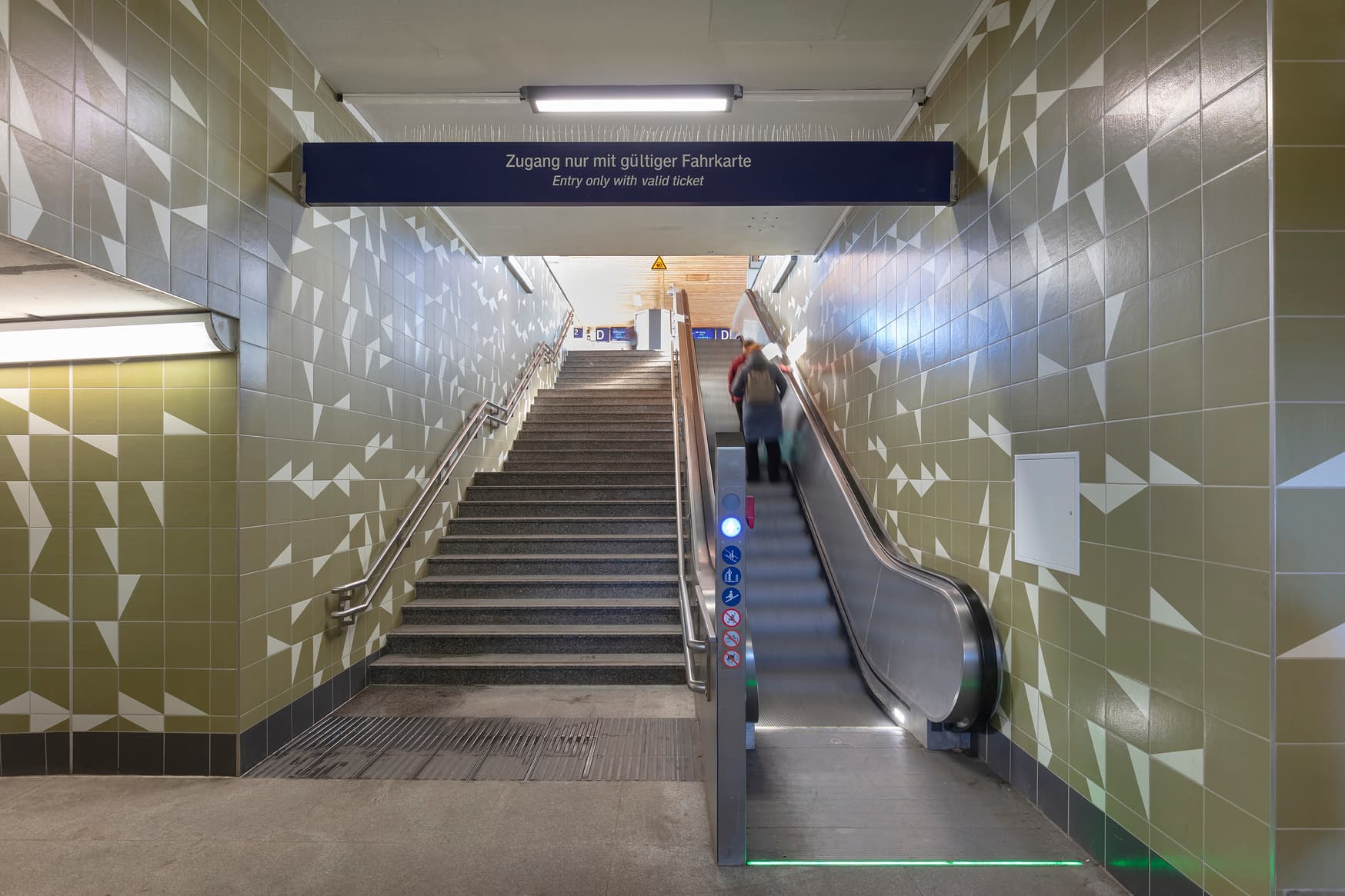 Treppenaufgang mit grün-weißer Fliesenmosaik, Stufenmarkierung und Handlauf.