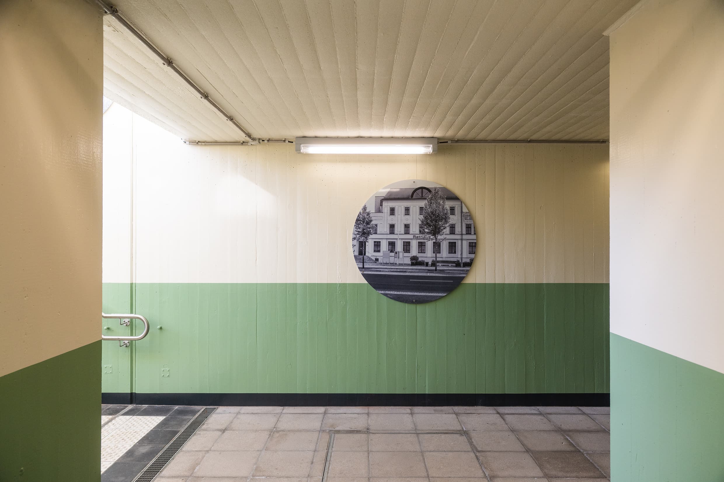 Blick auf eine Wand in der Personenunterführung: obere Hälfte wurde hellbeige, untere Hälfte grün gestrichen. Man sieht ein einzelnes, rundes Board in Schwarz/weiß, was ein Haus aus der Region darstellt.