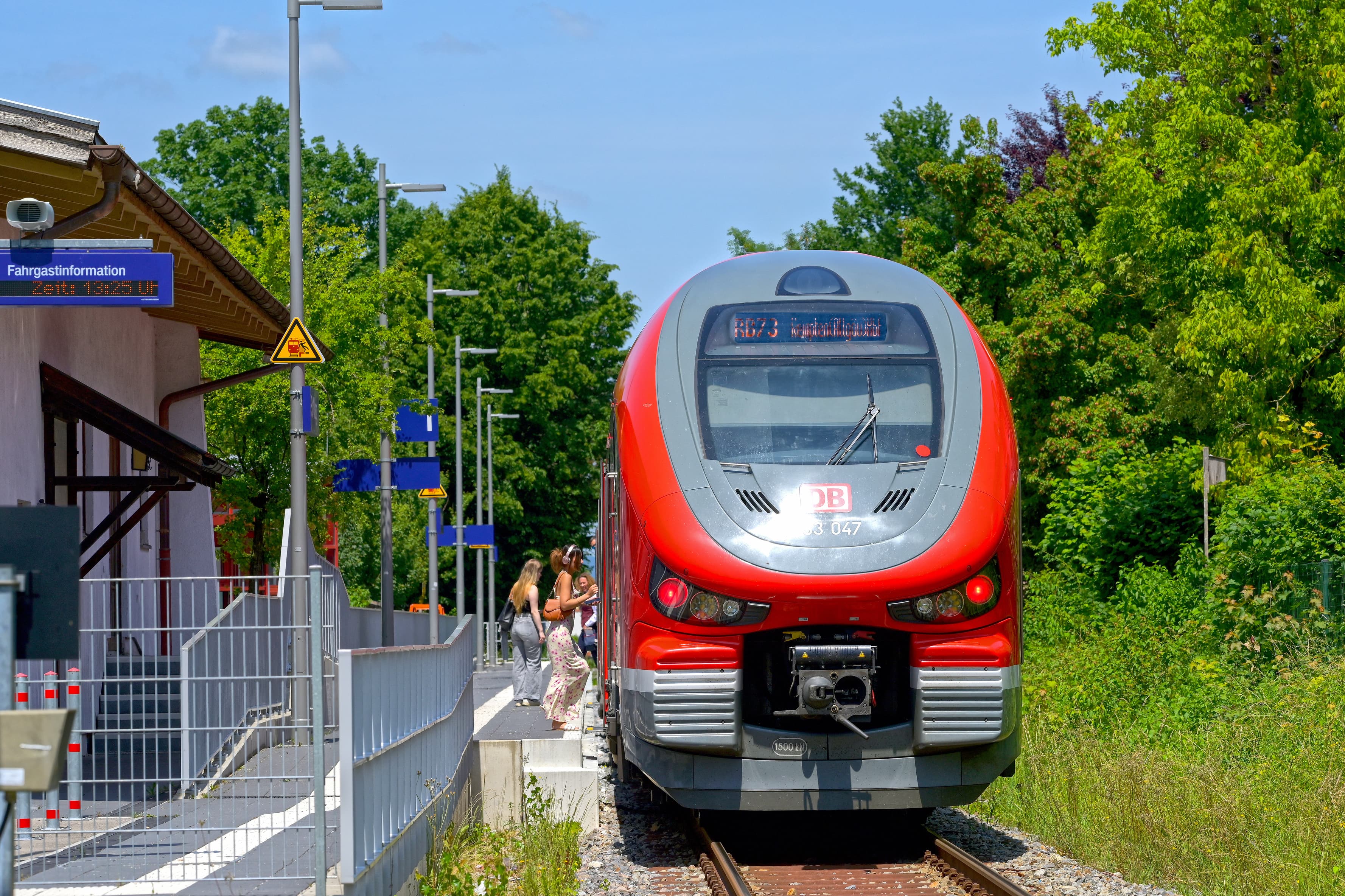 Ein haltender Zug am Bahnsteig. Es steiegen Reisende in den Zug. Am Anfang des Bahnsteiges hängt ein einzeiliger Informationsmonitor. 