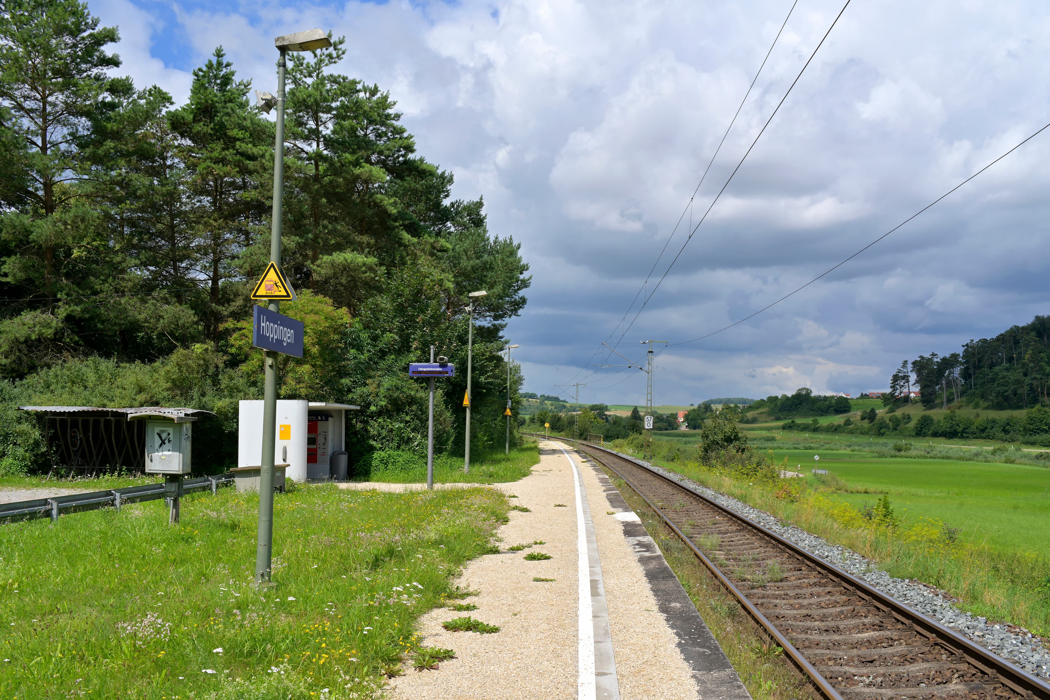 Links blühende Grünflächen. In der Mitte ein unebener mit Unkraut bewachsener Bahnsteig. Im Hintergrund ein Wetterschutzhaus aus Beton. Rechts im Bild verlaufen die Gleise. 