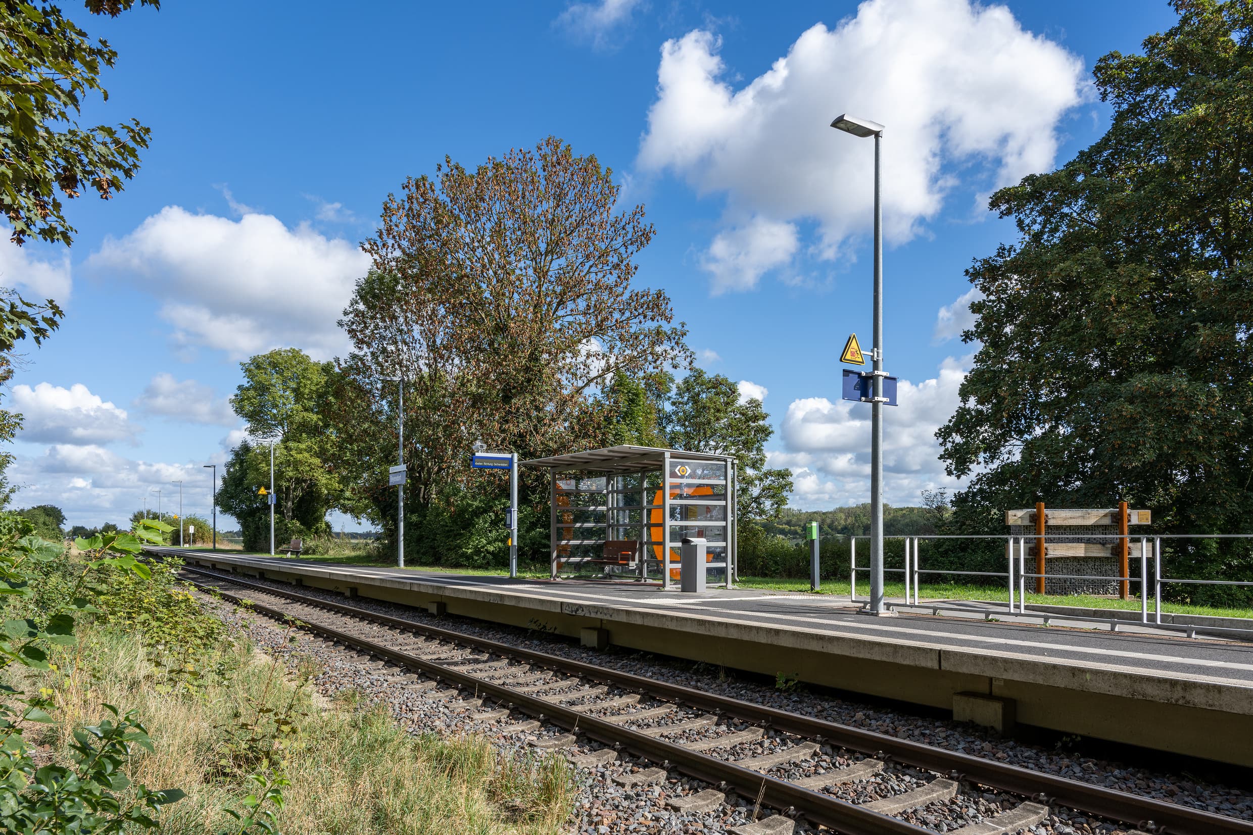 Ein eingleisiger Bahnsteig mit einem volltransparenten Wetterschutzhaus inkl. Vitrine und digitaler Reisendeninformation. Im Hintergrund ist noch das große Insektenhabitat zu sehen.