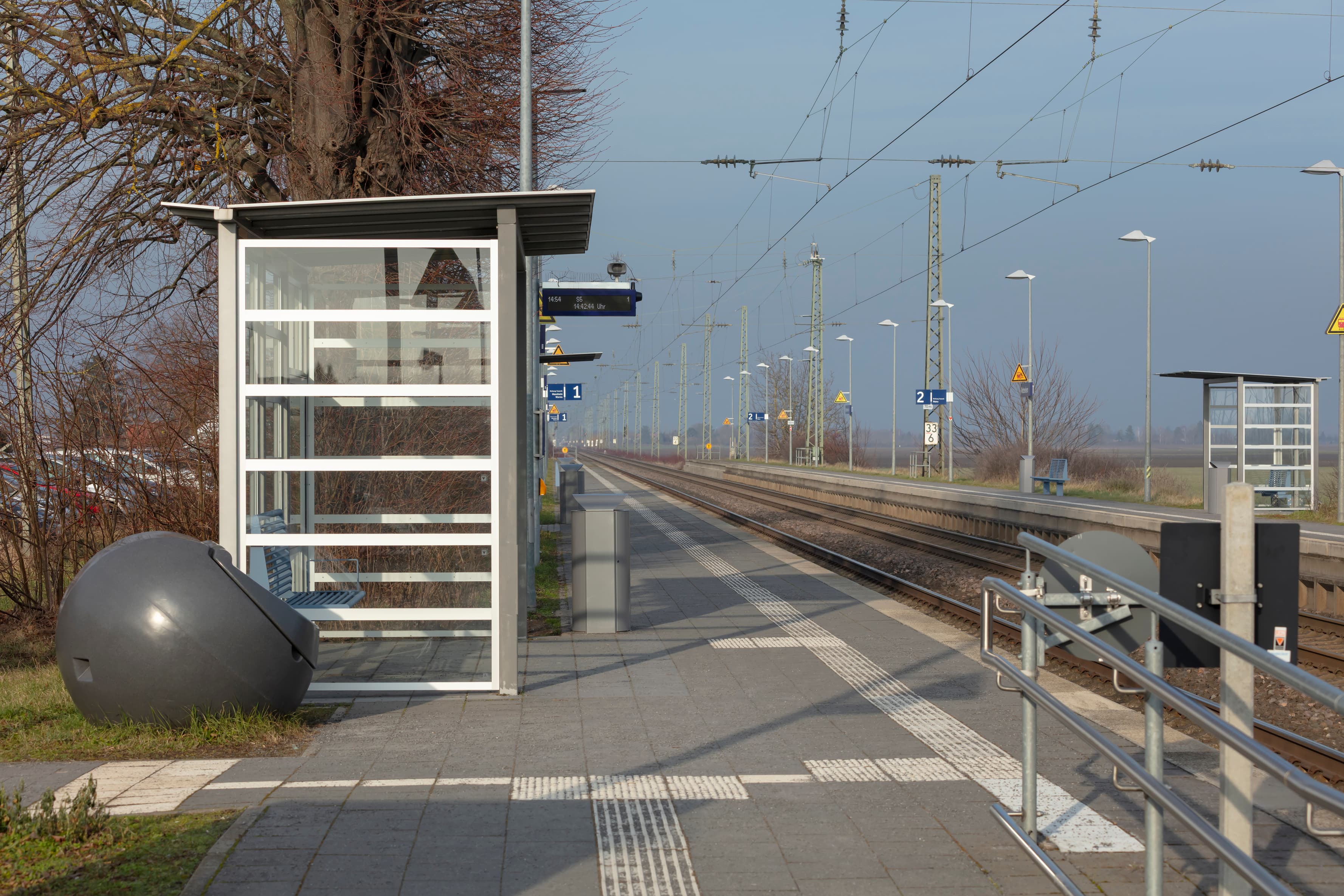 Beide Bahnsteige in voller Länge mit Wetterschutzhaus, blauen Sitzbänken, Mülleimern und Lampen, dazwischen die Gleise.