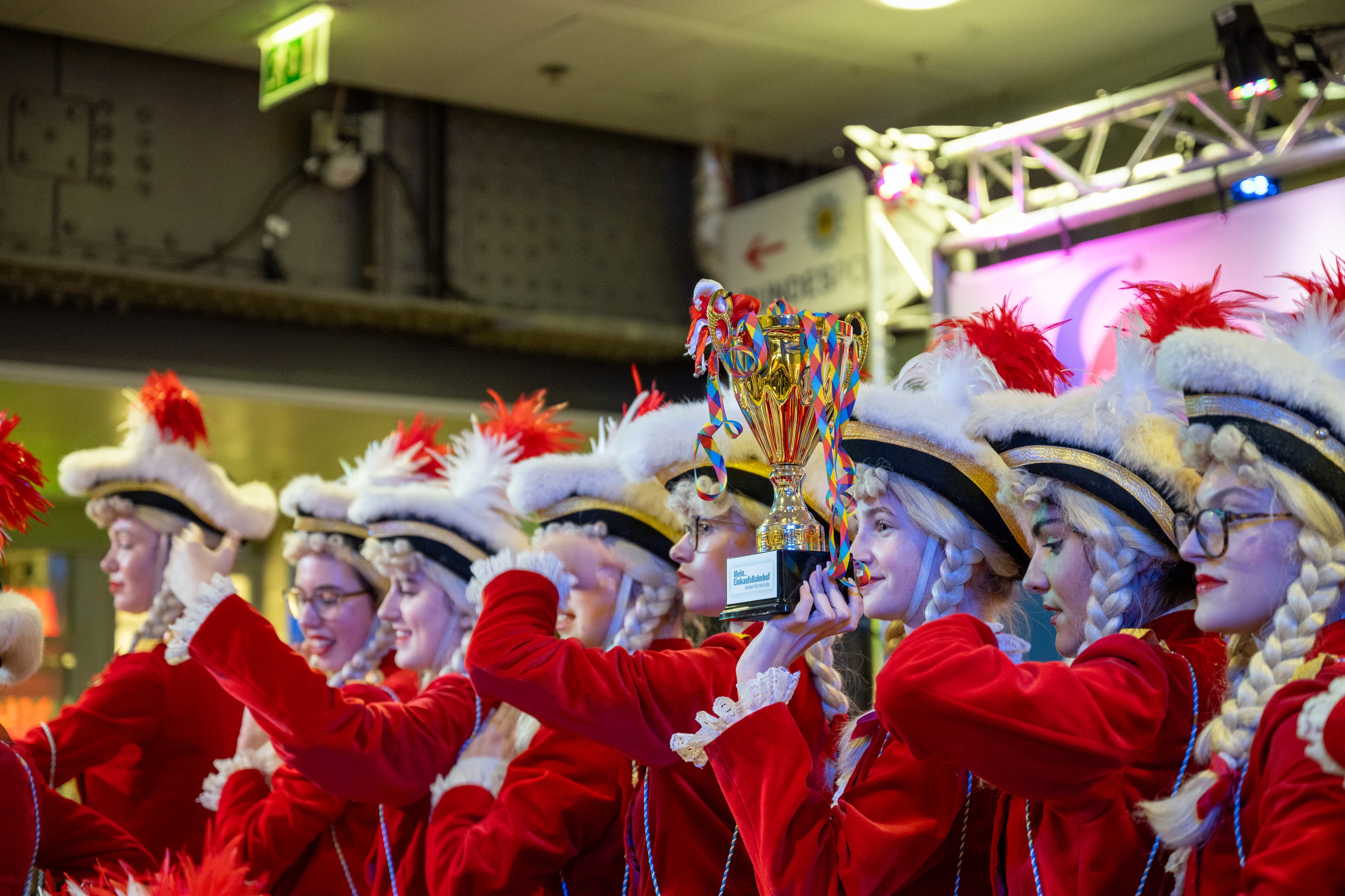 Gruppe von Mädchen in roten Tanz-Uniformen und weißen Federhüten hebt gemeinsam einen goldenen Pokal mit bunten Bändern in einer festlich beleuchteten Bahnhofshalle in die Höhe.