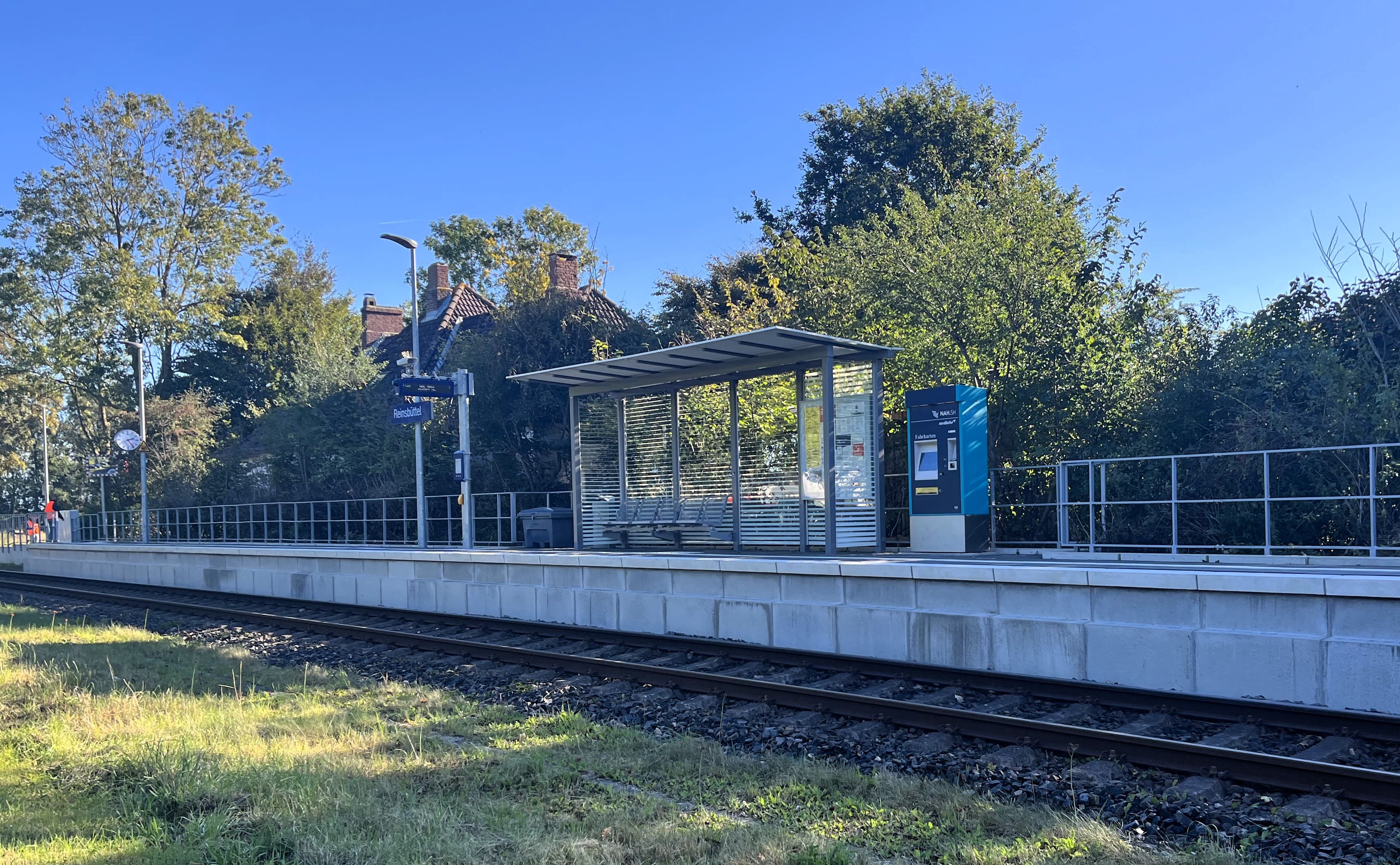 Auf dem langgestreckten neuen Bahnsteig steht ein Wetterschutzhaus, ein Fahrkartenautomat, Zugzielanzeiger und Bahnhofsuhr.