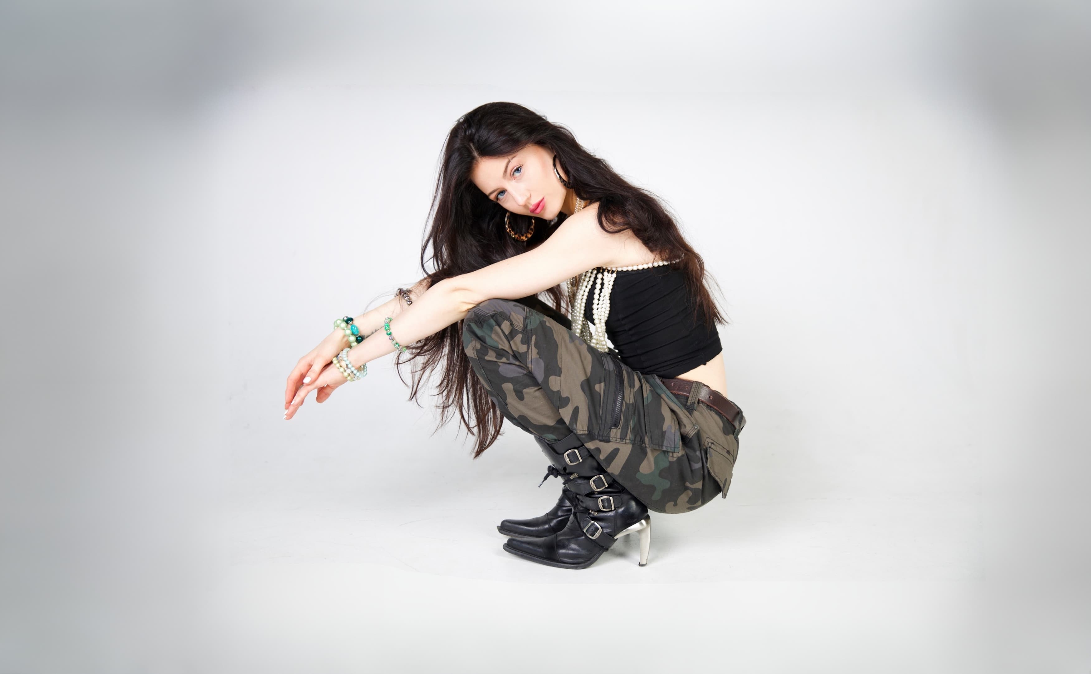 Nina Louisa mit langem Haar in Tarnhose, schwarzem Top und Stiefeln hockt mit ausgestreckten Armen vor weißem Hintergrund