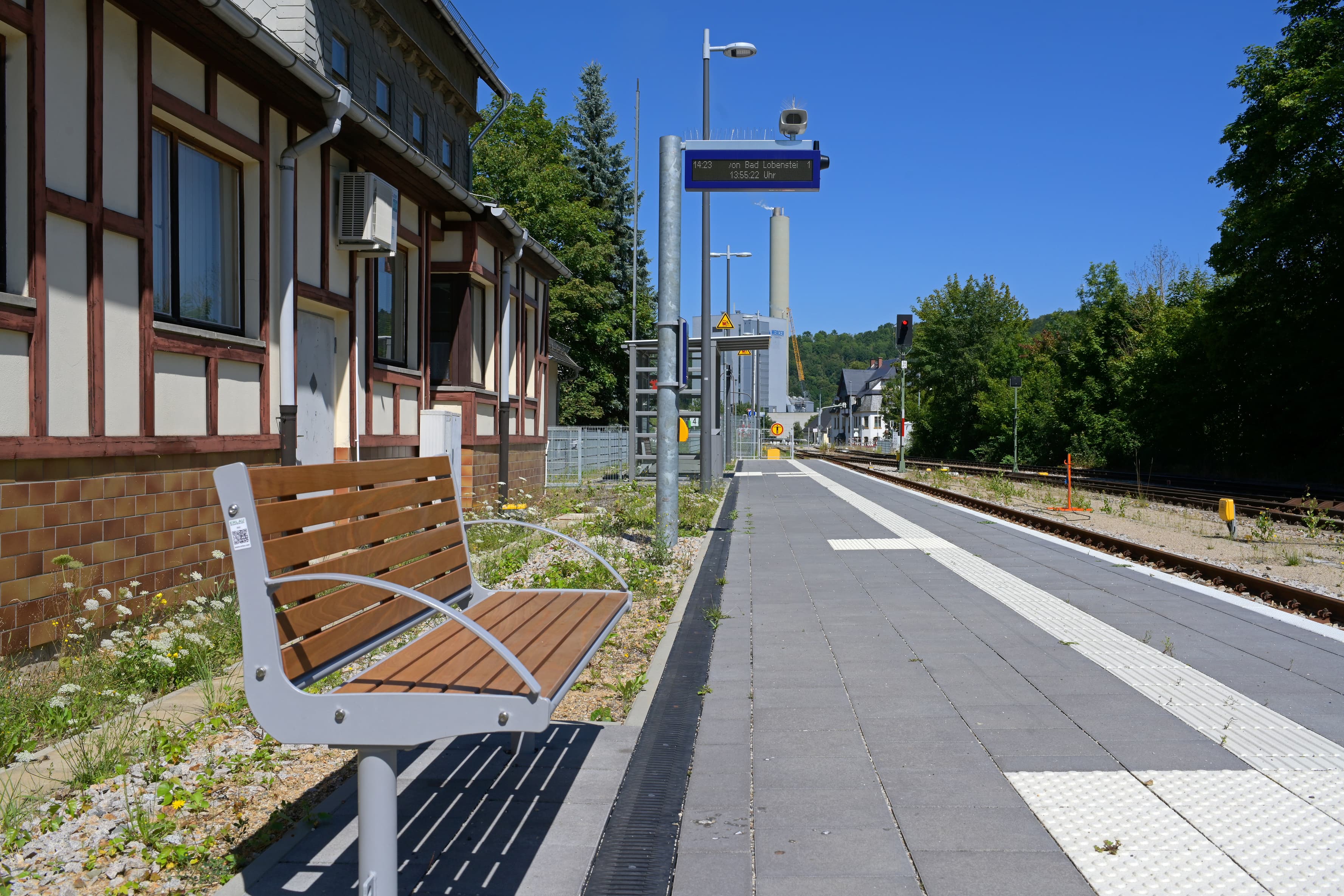 Blick auf den fertig umgebauten Bahnsteig mit holzbelatteter Sitzbank und zweizeiliger, digitaler Reisendeninformation.