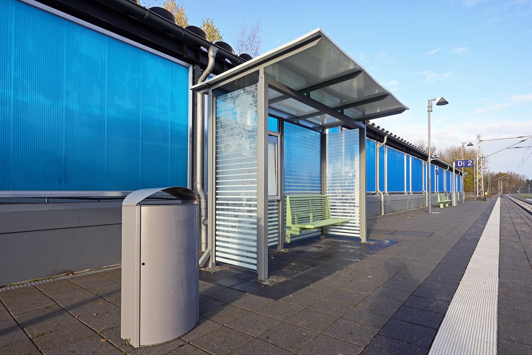 Ein Mülleimer, ein Wetterschutzhäuschen auf dem Bahnsteig mit taktilem Leitsystem. Dahinterliegende Wand ist blau.