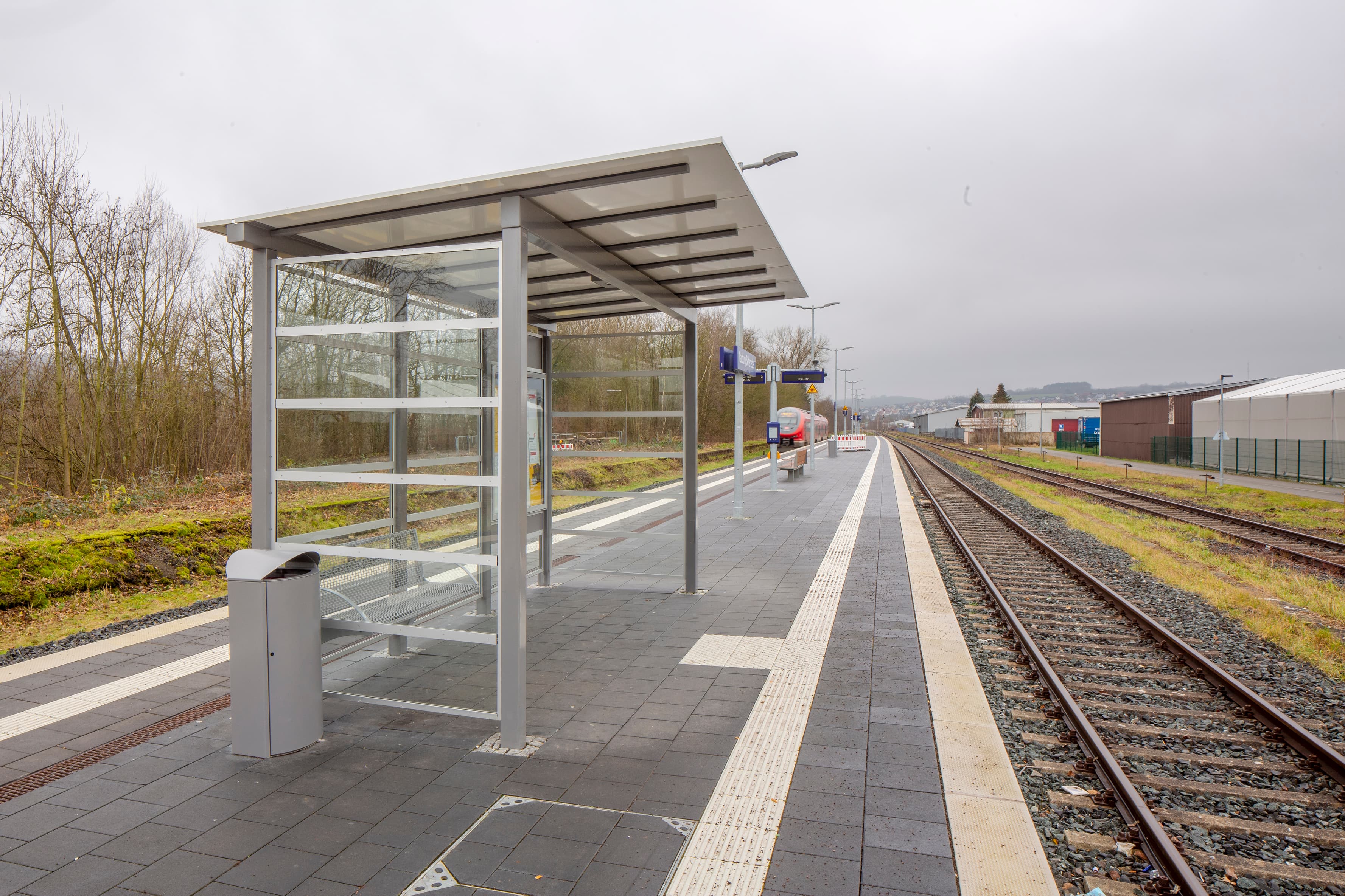 Wetterschutzhäuschen auf einem Bahnsteig, daneben ein Metallmülleimer.