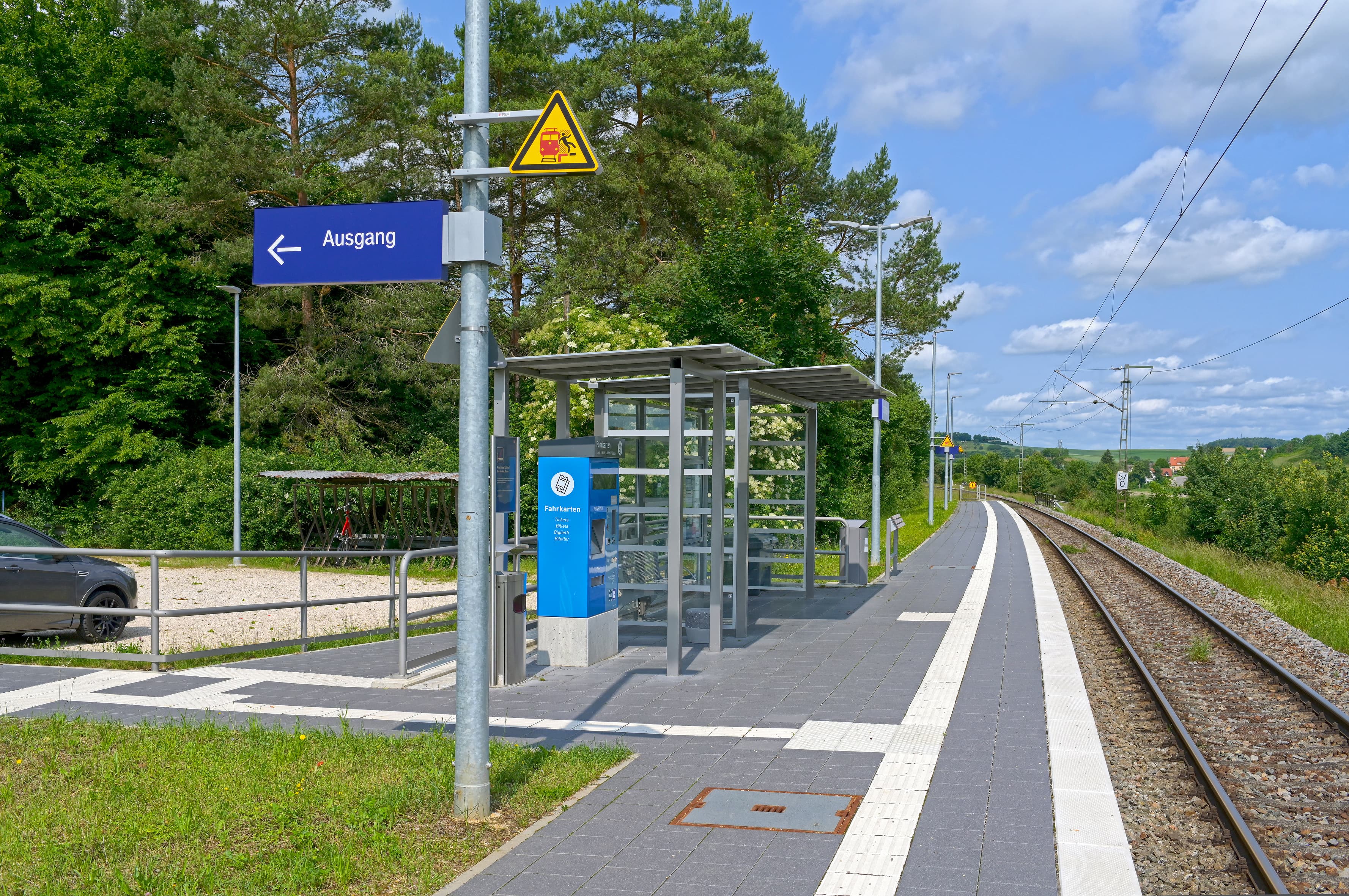 In der Bildmitte ein moderner Bahnsteig mit taktilem Leitsystem. Links im Bild eine Zugangsrampe sowie ein Wetterschutzhaus. Rechts verlaufen die Gleise. 