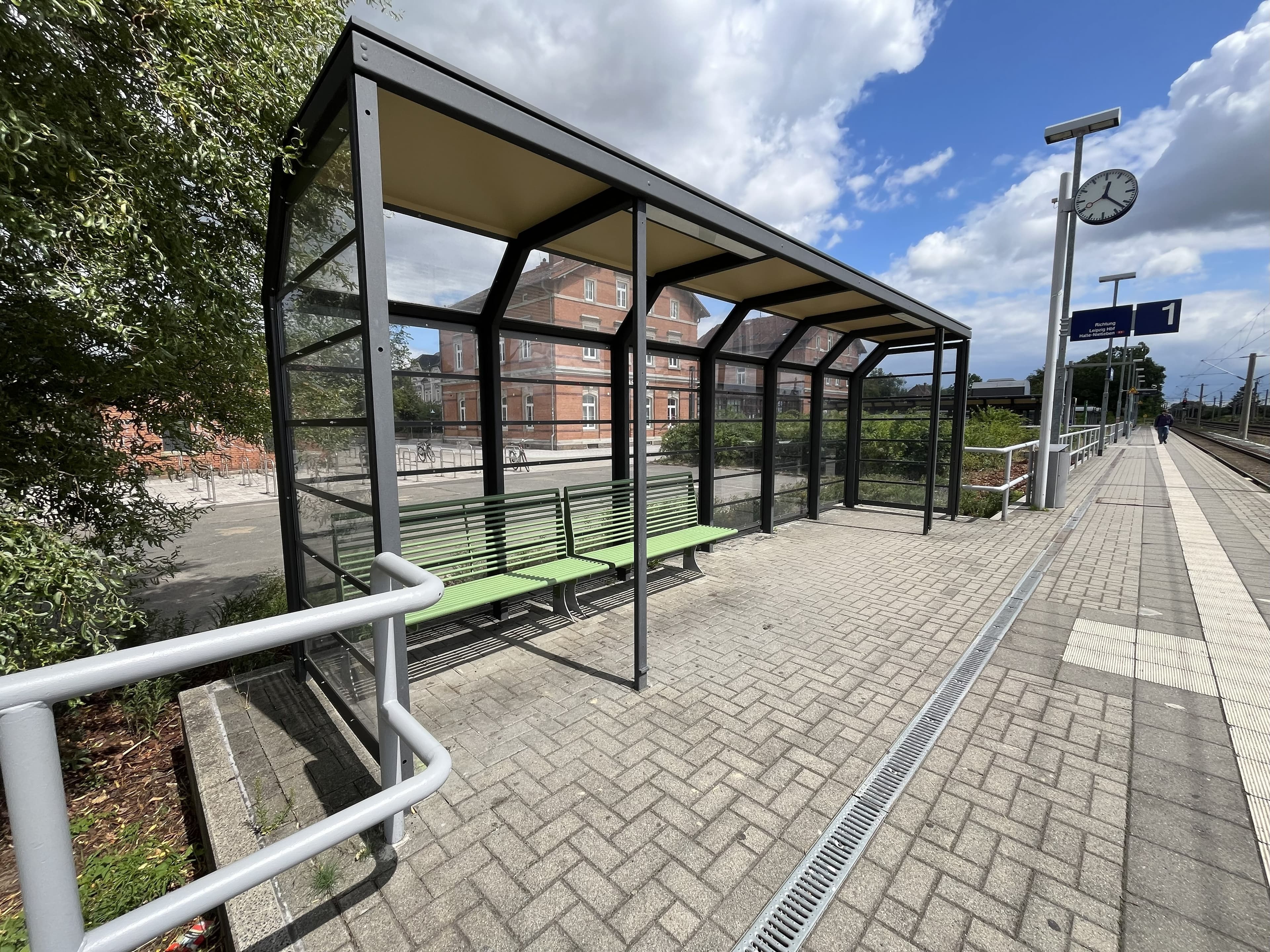 Zu sehen ist eines der neuen Wetterschutzhäuser mit transparenten Wandelementen und grünen Sitzbänken. 