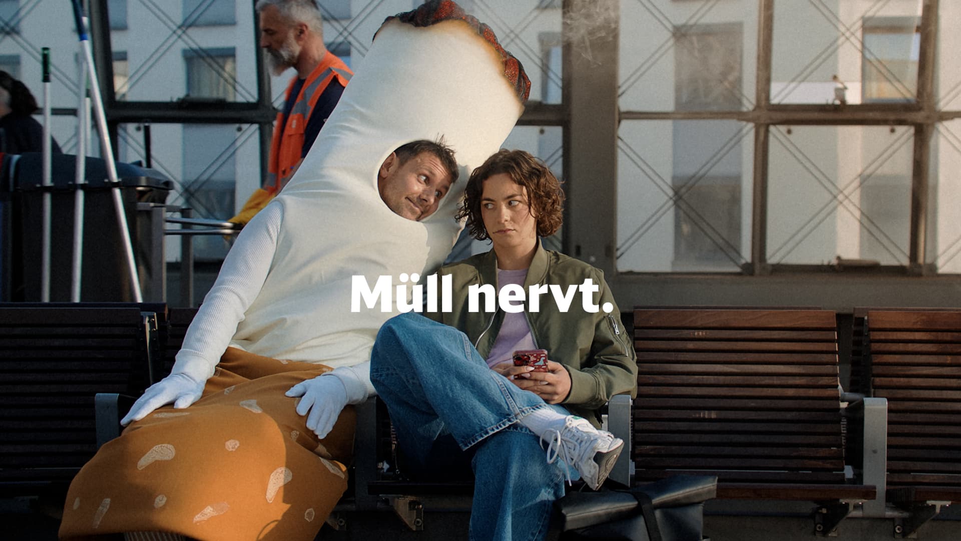 A person disguised as a cigarette sits on a bench on a platform next to a passenger and tries to read along on her mobile phone. The words 