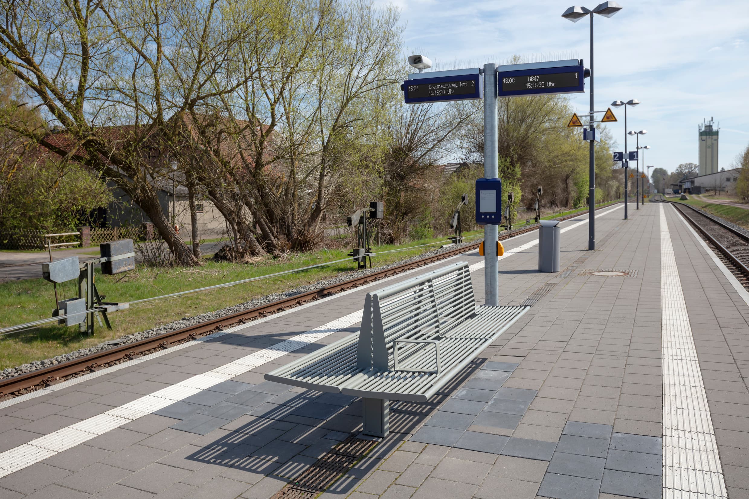 Moderne Sitzbank und ein Mast mit Zuginformationen am Bahnsteig.