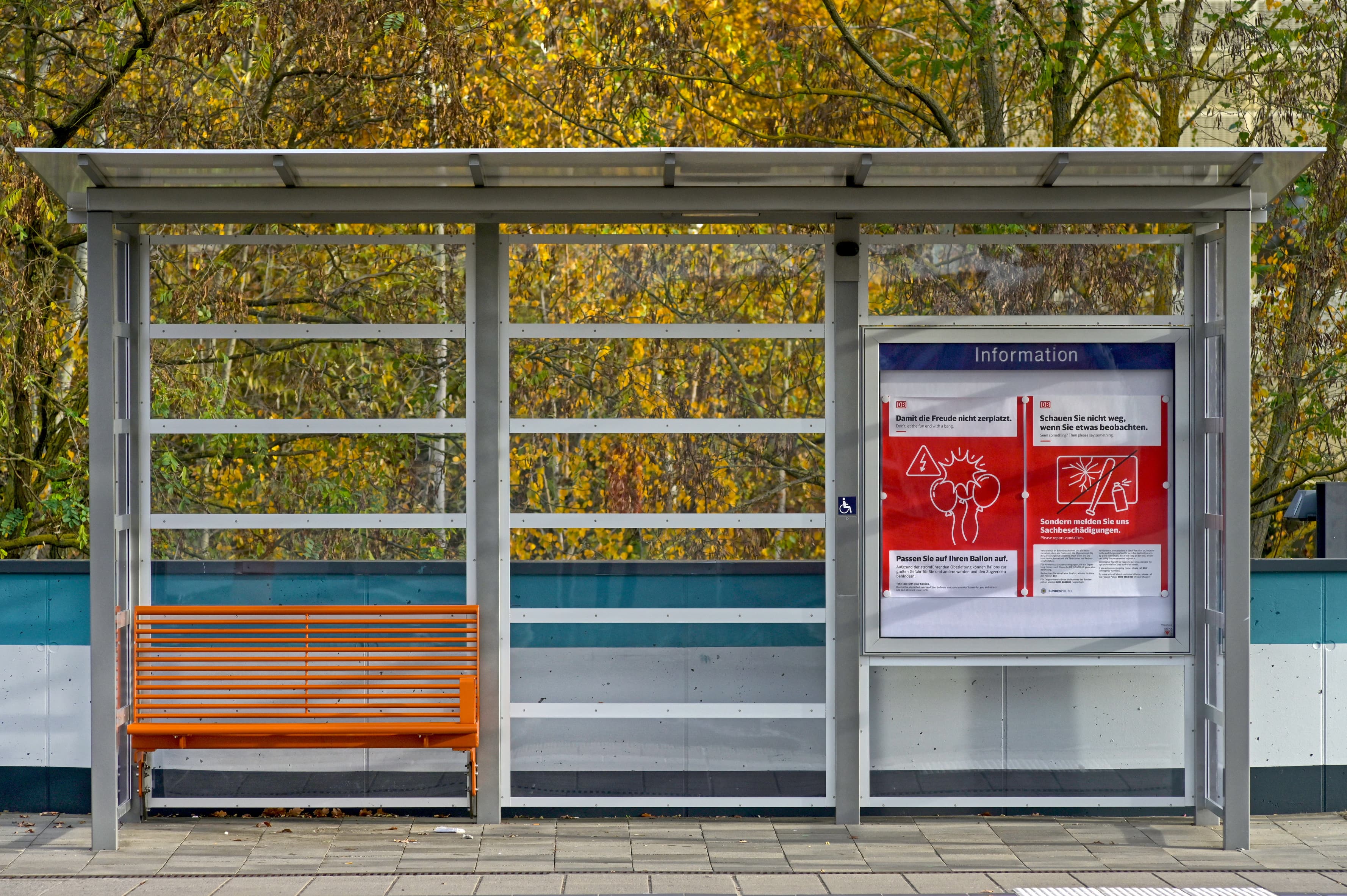 Blick auf ein neues Wetterschutzhaus. Die Wände sind von oben bis unten verglast. Darin steht eine neue Bank im satten Orange des neuen Farbkonzepts.