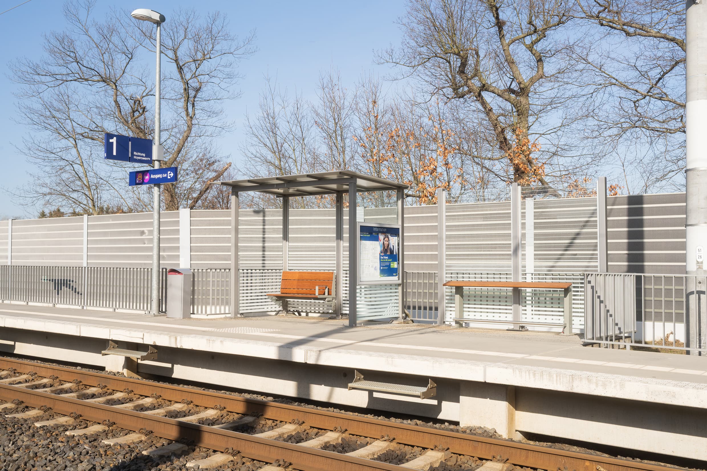 Voll transparentes Wetterschutzhaus mit Holzbank. Rechts daneben steht ein Anlehner aus Holz, links daneben der Mülleimer. Im Hintergrund sieht man die graue Lärmschutzwand.