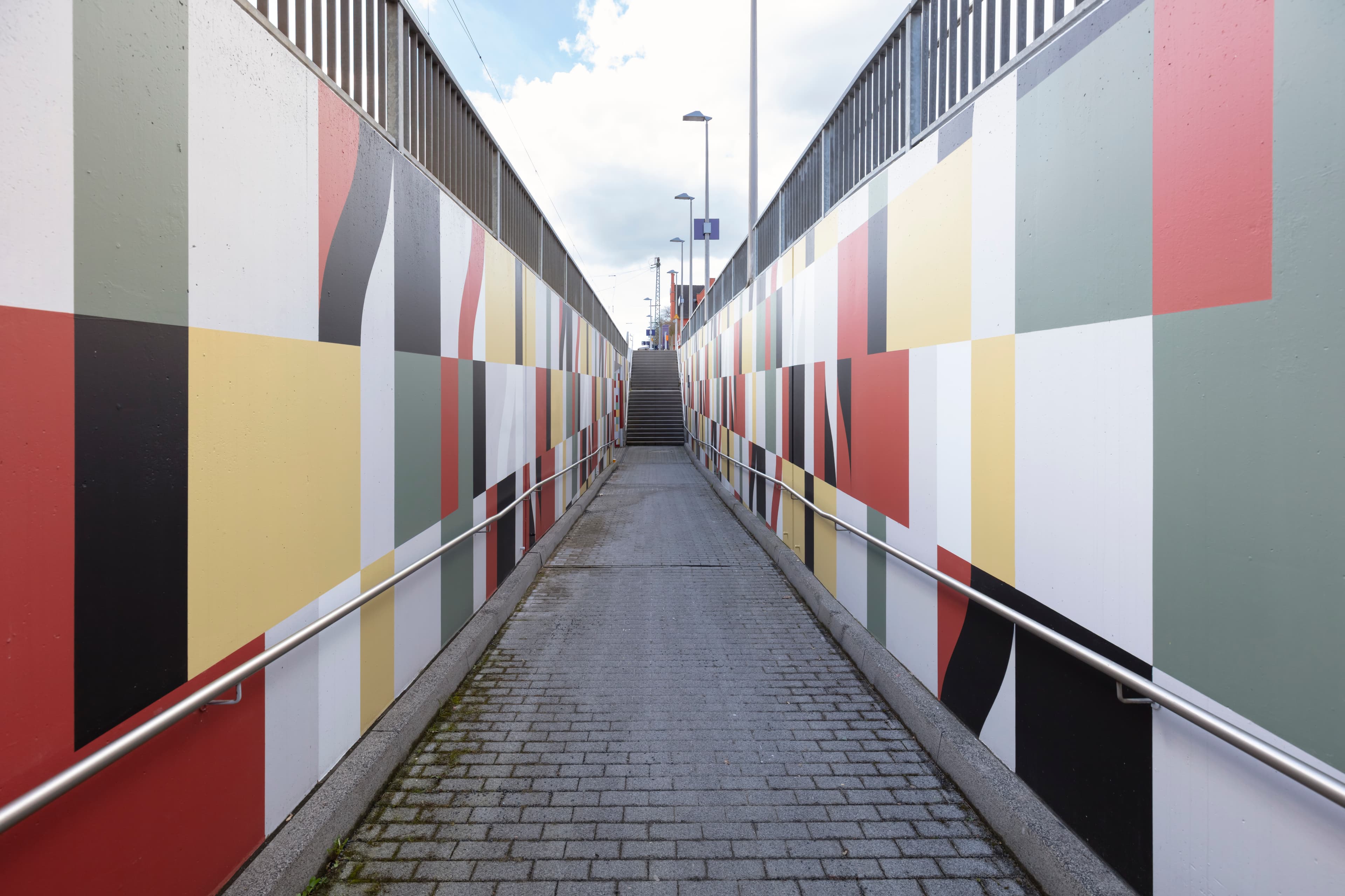 Blick auf den Aufgang einer bunt gefliesten Personenunterführung. Rechts und links davon Wände mit Handläufen.