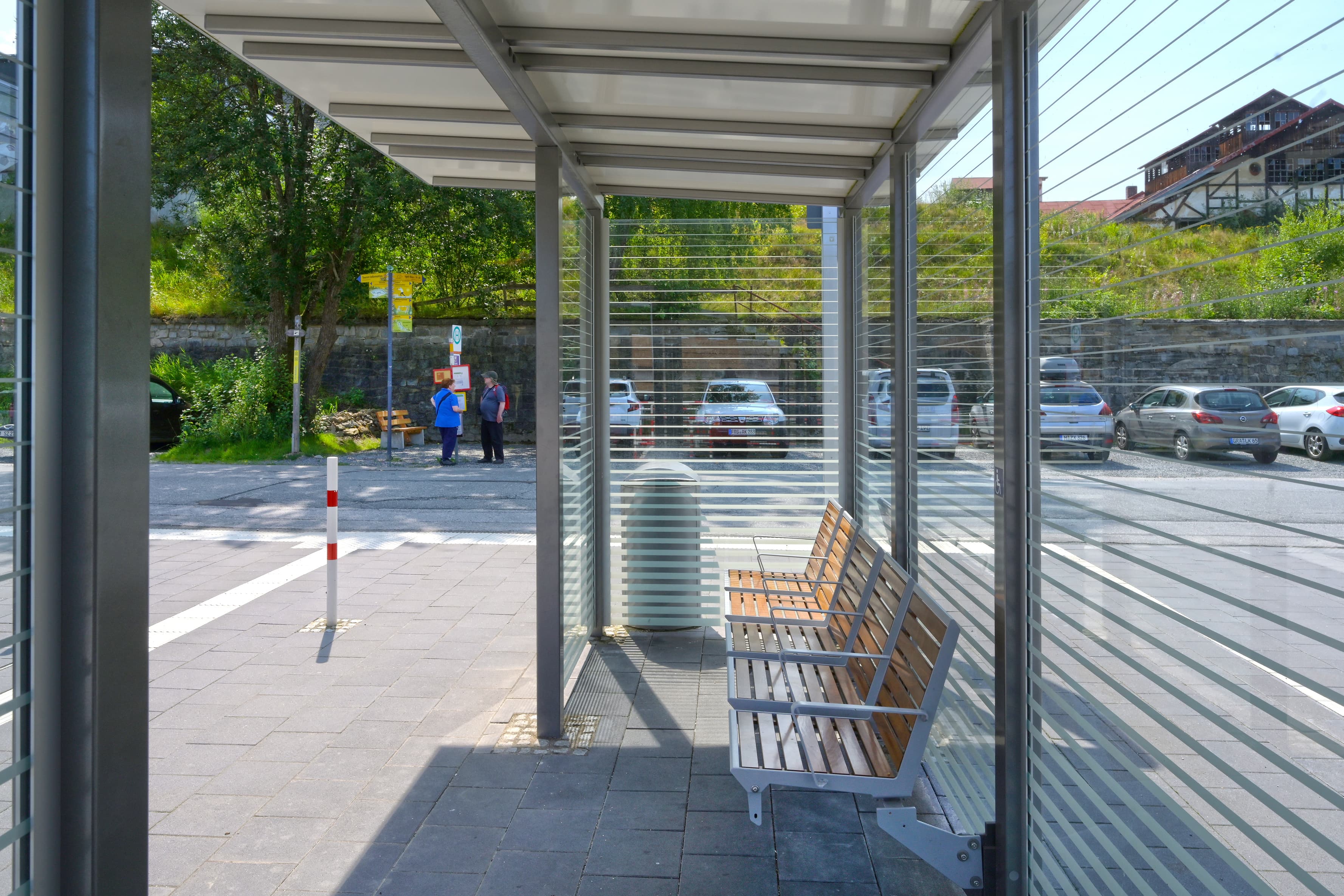 Das neue und vollverglaste Wetterschutzhaus mit Sitzgelegenheit auf dem Weg zu den Gleisen. Dahinter eine Straße. Links im Bild das taktile Leitsystem.