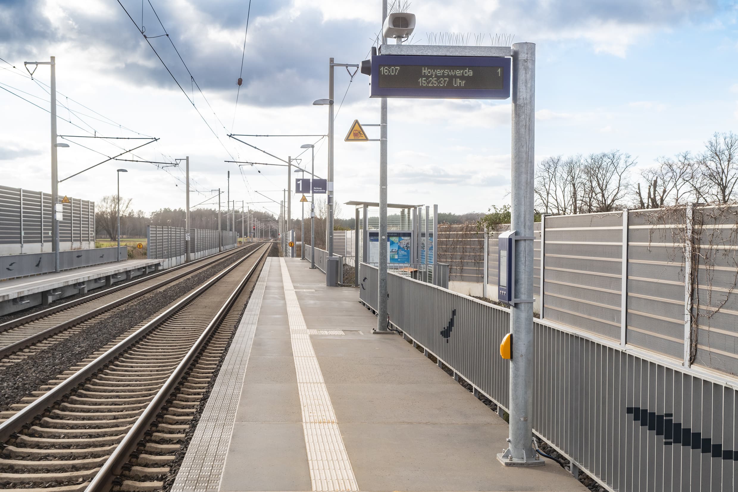 Zweizeiliger, digitaler Anzeiger am Mast. Darunter wurde der digitale Bauanzeiger angebracht, der baubedingte Fahrplanänderungen anzeigt. Im Hintergrund sieht man noch das Wetterschutzhaus.