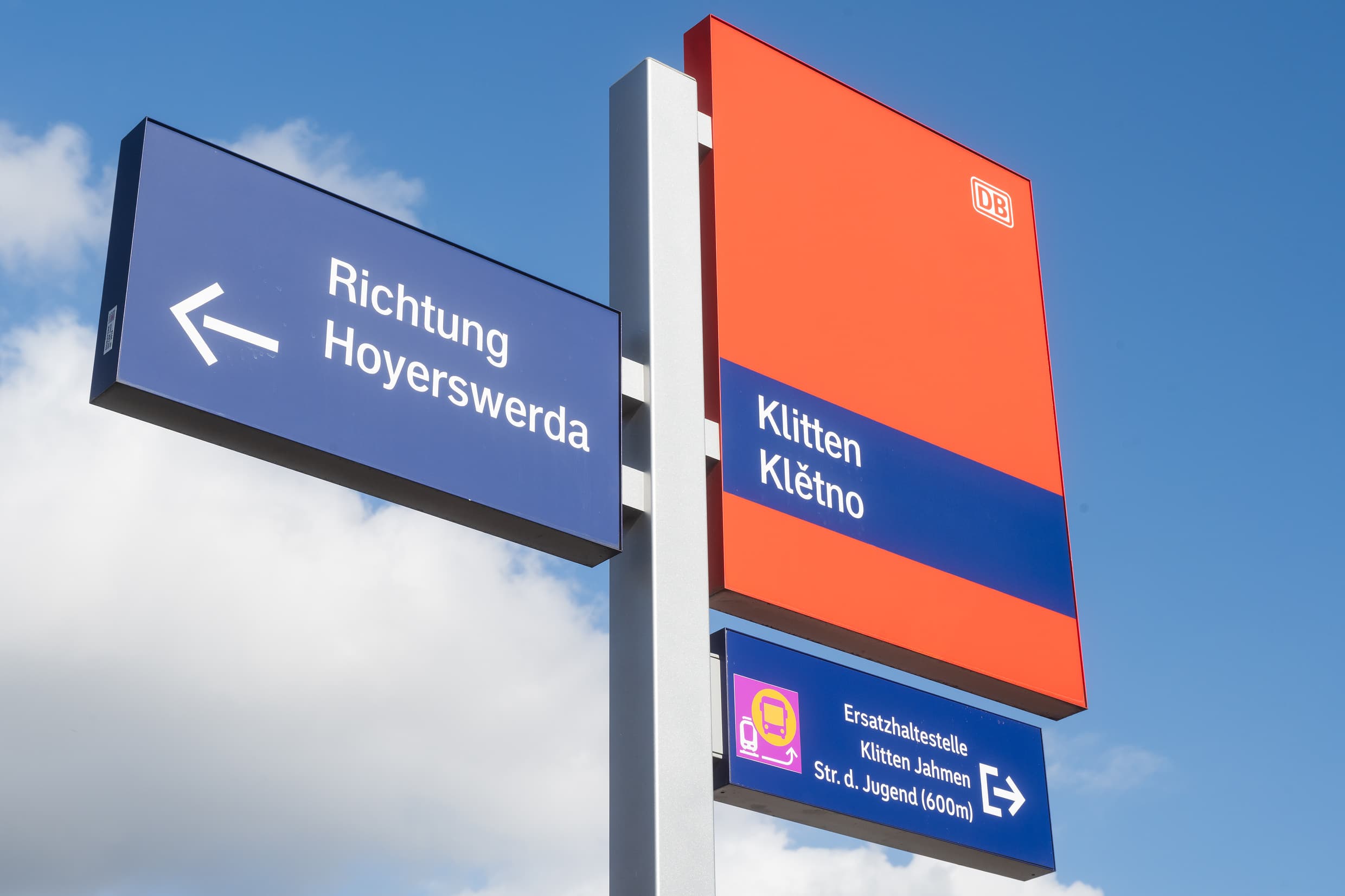 Stationsfahne mit rotem Schild und dem Stationsnamen Klitten auf deutsch und sorbisch, zusätzliche Orientierung durch Pfeile in Richtung Ersatzverkehr und Zugrichtung.