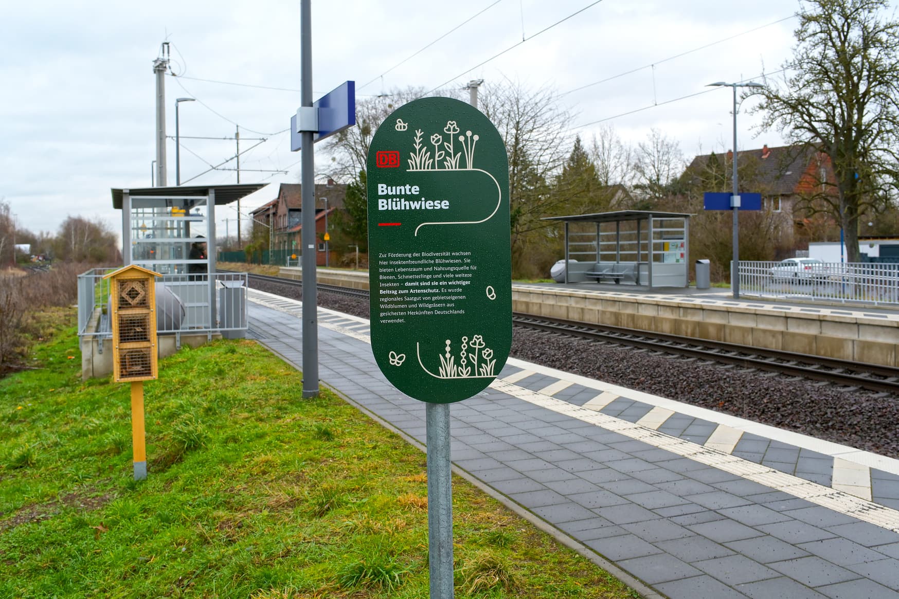 Hinweisschild zur Blühwiese neben dem Bahnsteig, im Hintergrund ein Insektenhotel und zwei Bahnsteige mit je einem Bahnhofsschild und einem verglasten Wetterschutzhäuschen.