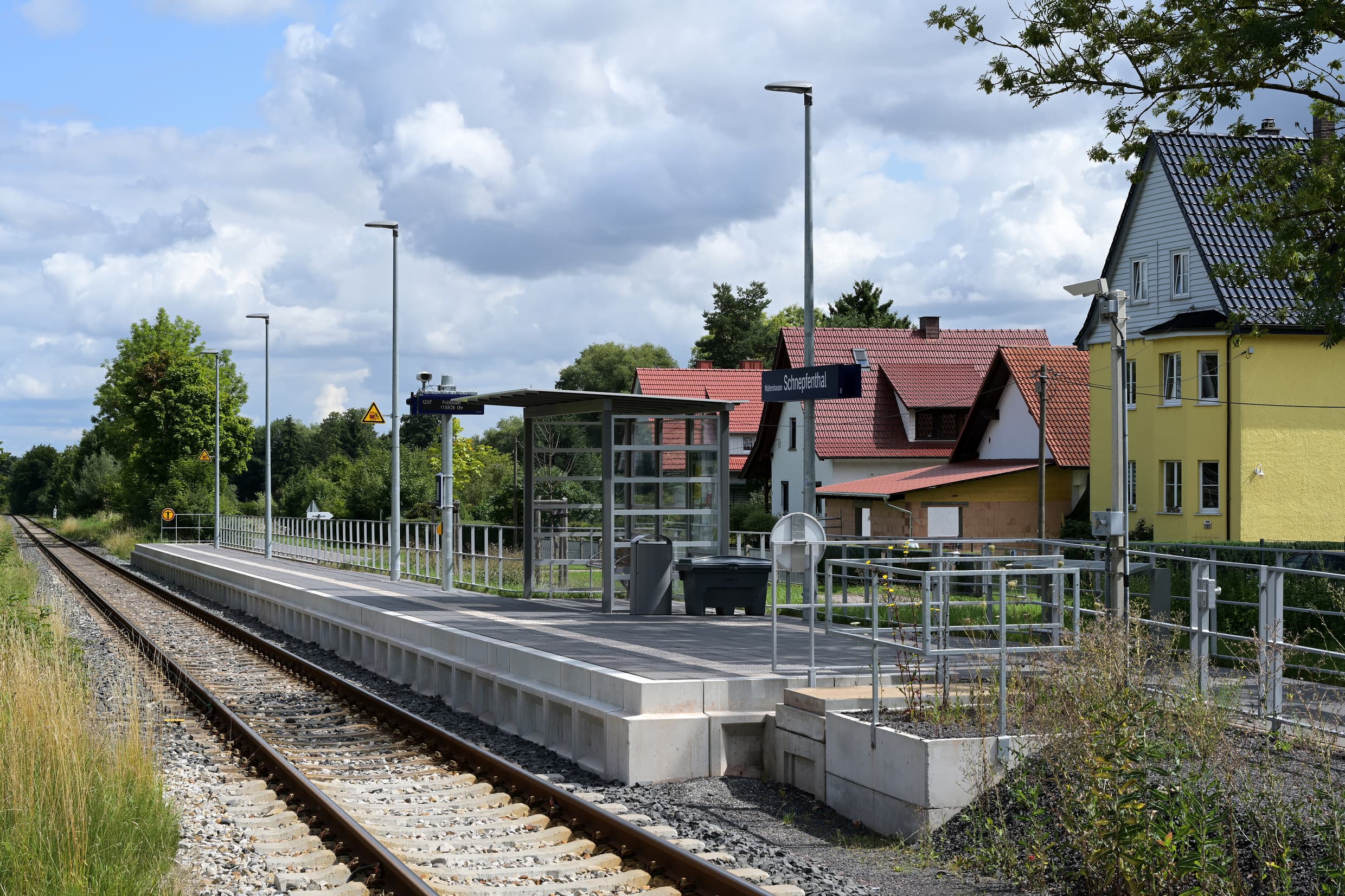 Man sieht den einseitigen moderne Bahnsteig, ein voll transparentes Wetterschutzhaus, ein digitaler Anzeiger und weitere Ausstattungselemente.