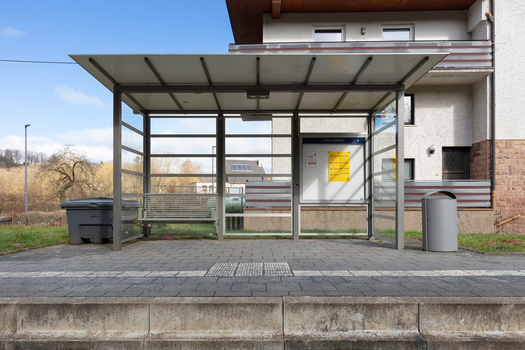 Ein transparentes Wetterschutzhaus mit Sitzbank und Vitrine. Daneben ein Mülleimer, vorne auf dem Bahnsteig das taktile Bodenleitsystem mit Aufmerksamkeitsfeld. 