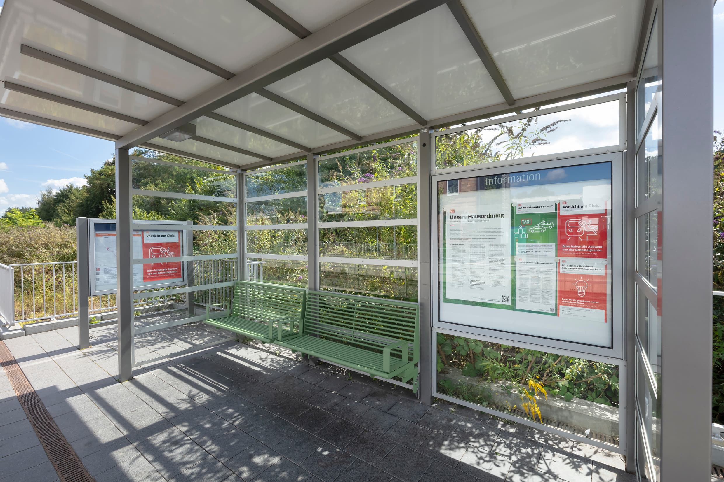 Grüne Sitzbänke in einem Wetterschutzhäuschen aus Verbundsicherheitsglas auf einem Bahnsteig.