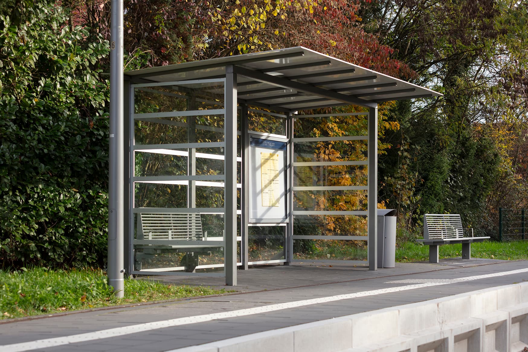 Transparentes Wetterschutzhaus auf dem Bahnsteig, daneben ein Mülleimer und eine Sitzbank.