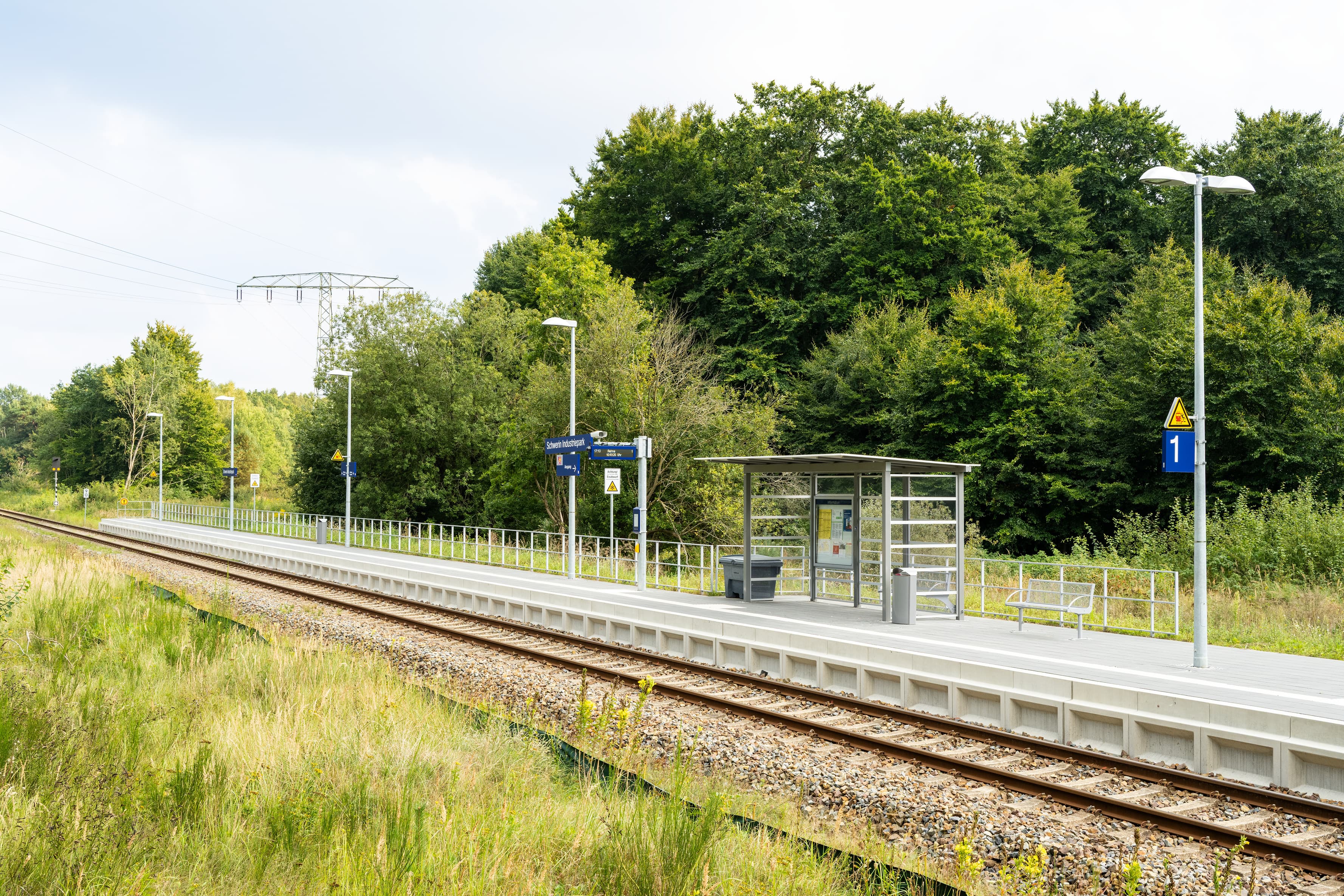 Blick über die Gleise auf den neuen Bahnhof Schwerin Industriepark. Darauf stehen das vollverglaste Wartehaus und die Reisendeninformationssysteme. Hinter dem Bahnhof ist die grüne Umgebung zu sehen. 