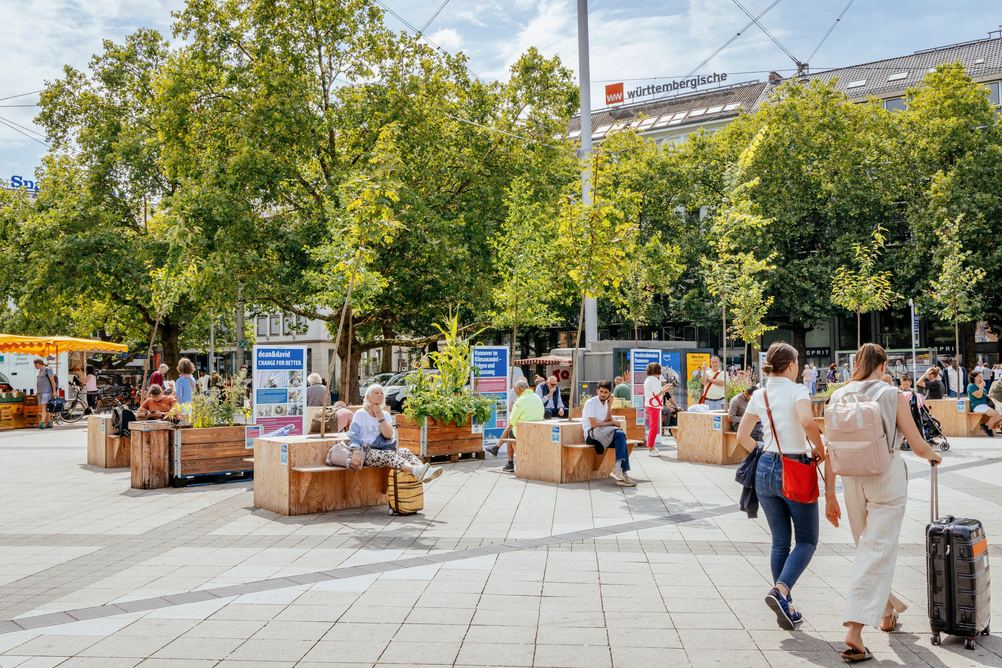Blick auf bepflanzte Sitzgelegenheiten auf dem Vorplatz des Hannover Hbf. Einige Menschen sitzen dort. Im Hintergrund ist ein Marktstand und einige Bäume zu erkennen. Im Vordergrund sieht man zwei Passantinnen mit Koffer.
