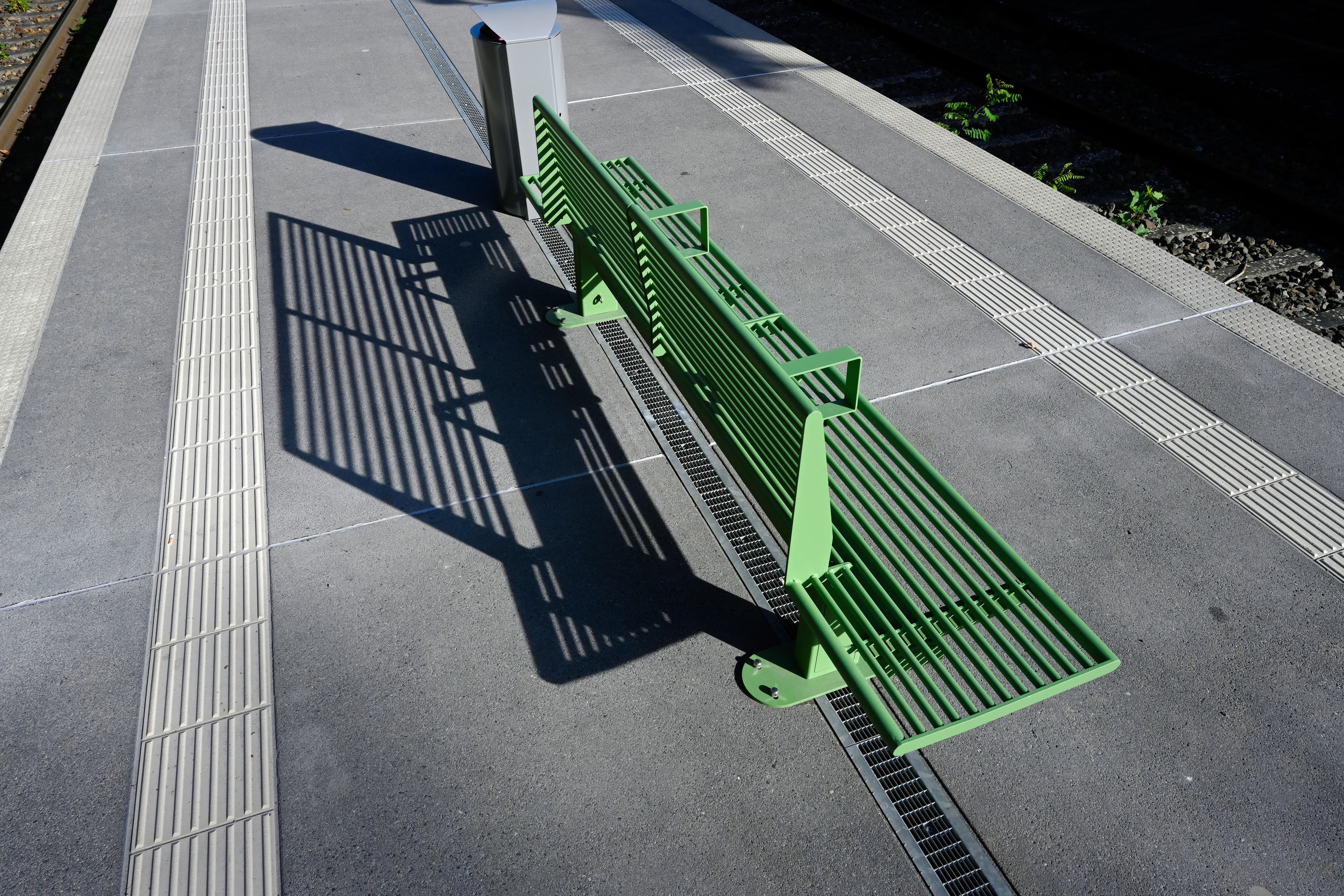 Eine grüne Sitzbank mit Rückenlehne auf einem Bahnsteig, rechts und links Blindenleitstreifen.