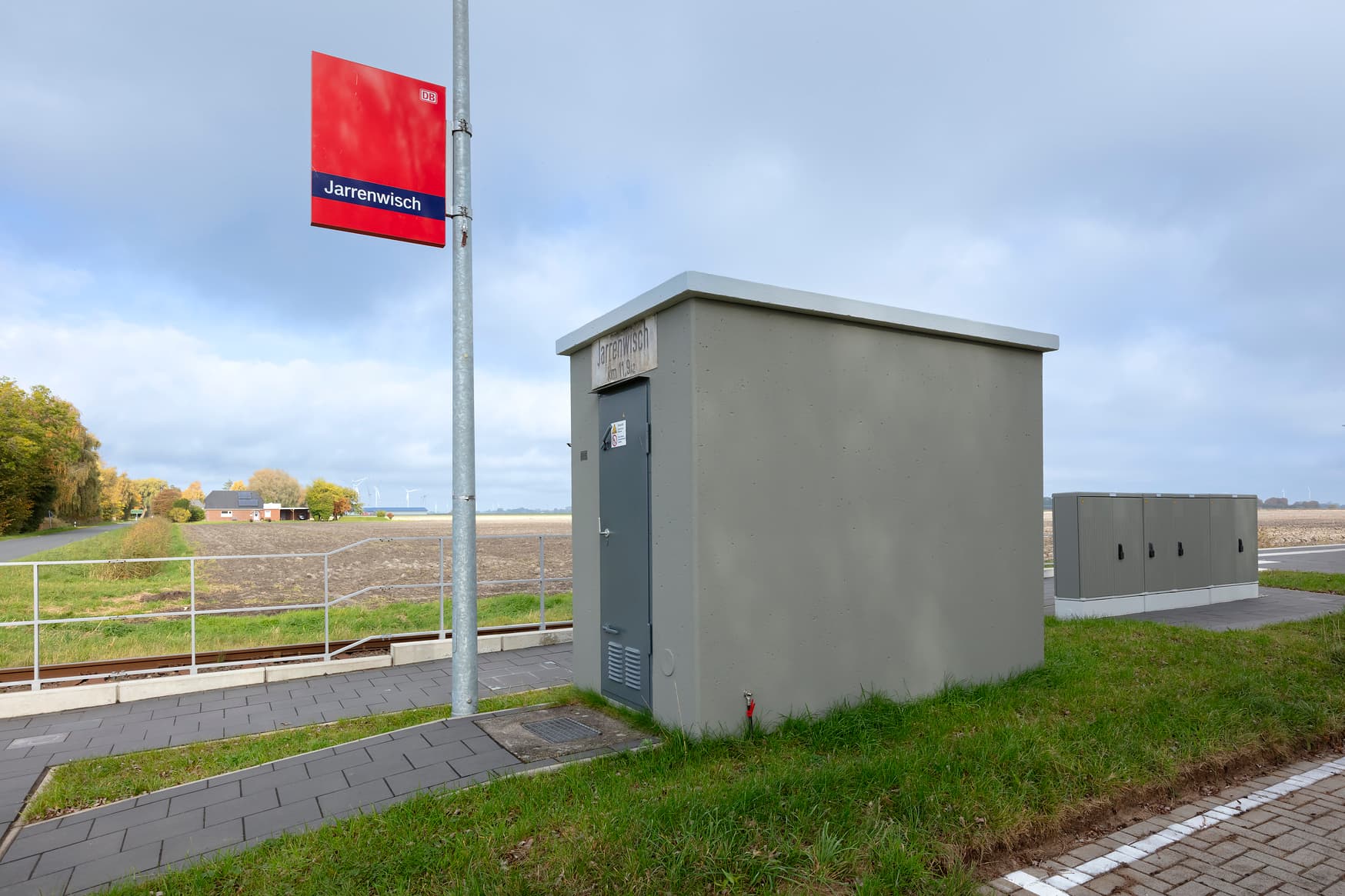 Bahnhofsschild in rot von Jarrenwisch daneben grün gestrichenes Trafohaus und grün gestrichene Verteilerkästen.