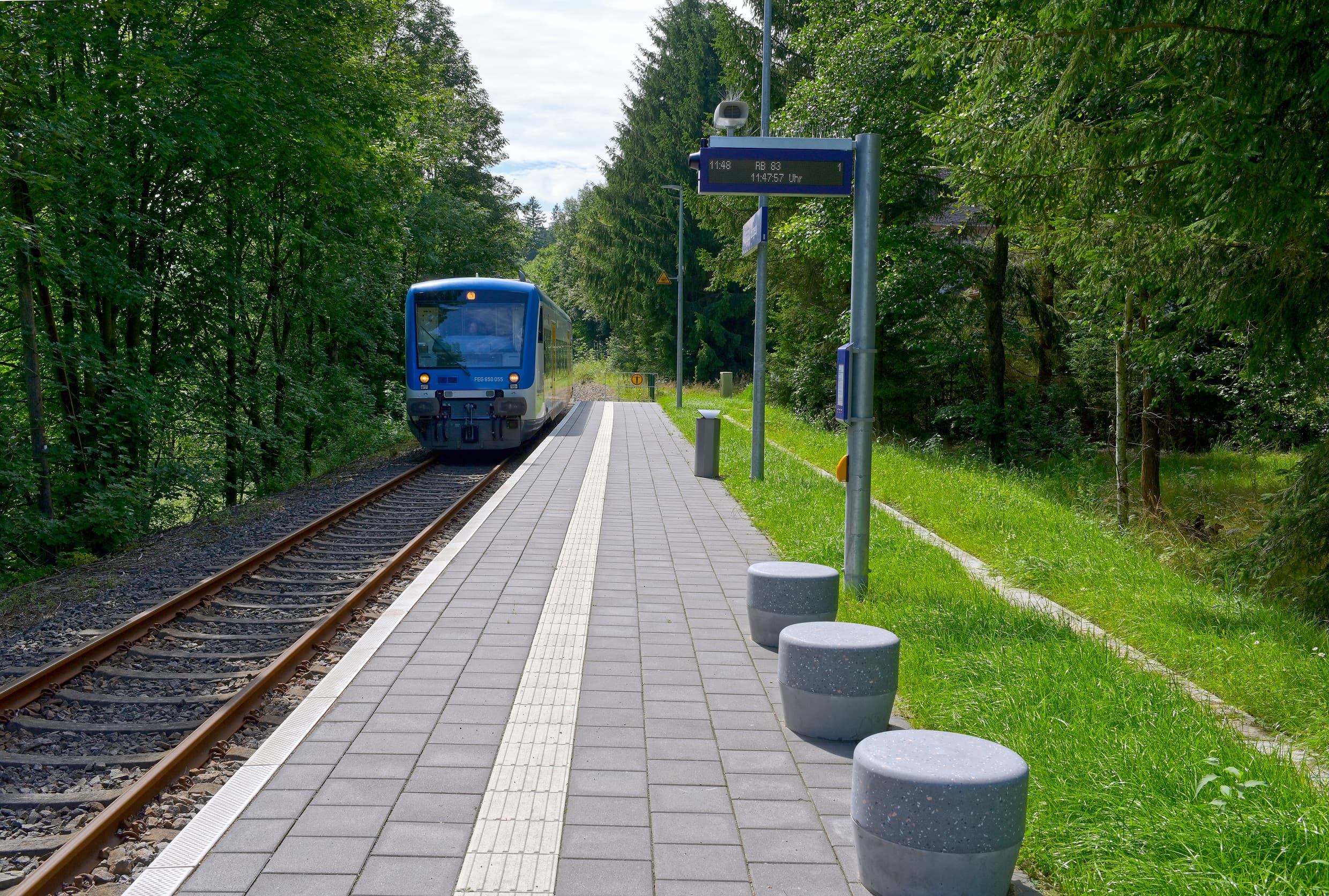 Einfahrender blauer Zug am neu gebauten, einseitigen Bahnsteig. Auf diesem sieht man drei Betonsitzhocker