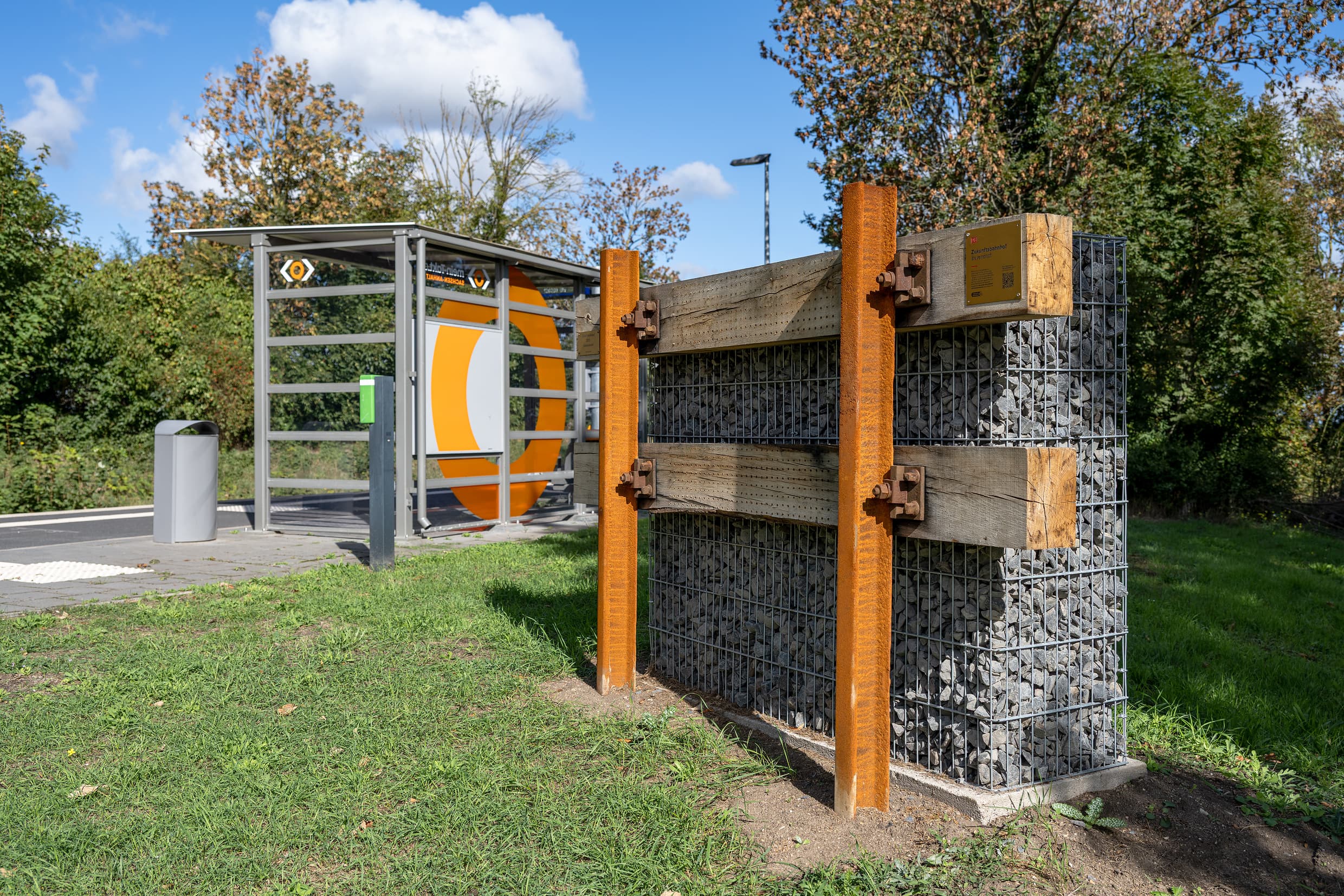 Im Vordergrund steht ein Insektenhotel in Form einer Gabionenwand mit angedeuteten Schienen als Gestaltungselement. Im Hintergrund ist das Wetterschutzhaus mit der aufgabtenträgerbezogenen orangenen Folierung zu sehen.