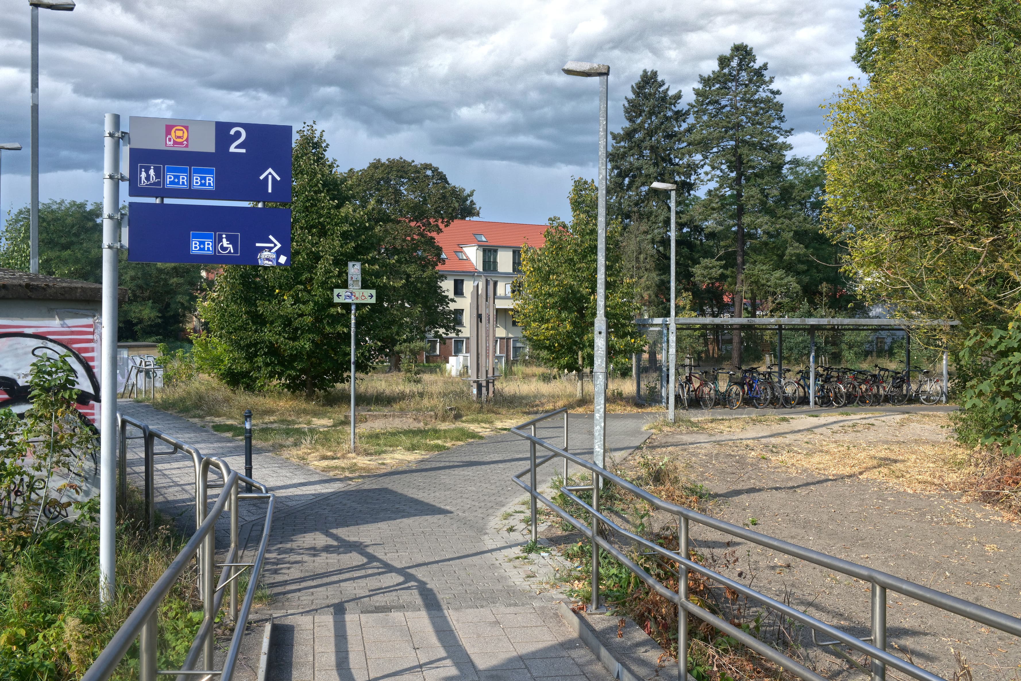 Ein blaues Schild, dass eine Zuwegung die nach rechts und links abführt, flaniert. Am rechten Weg sieht man Fahrradständer.