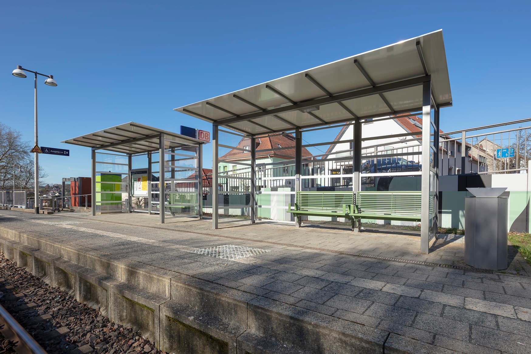 Zwei Wetterschutzhäuschen mit Sitzbänken auf einem Bahnsteig. Davor befindet sich das Gleis.