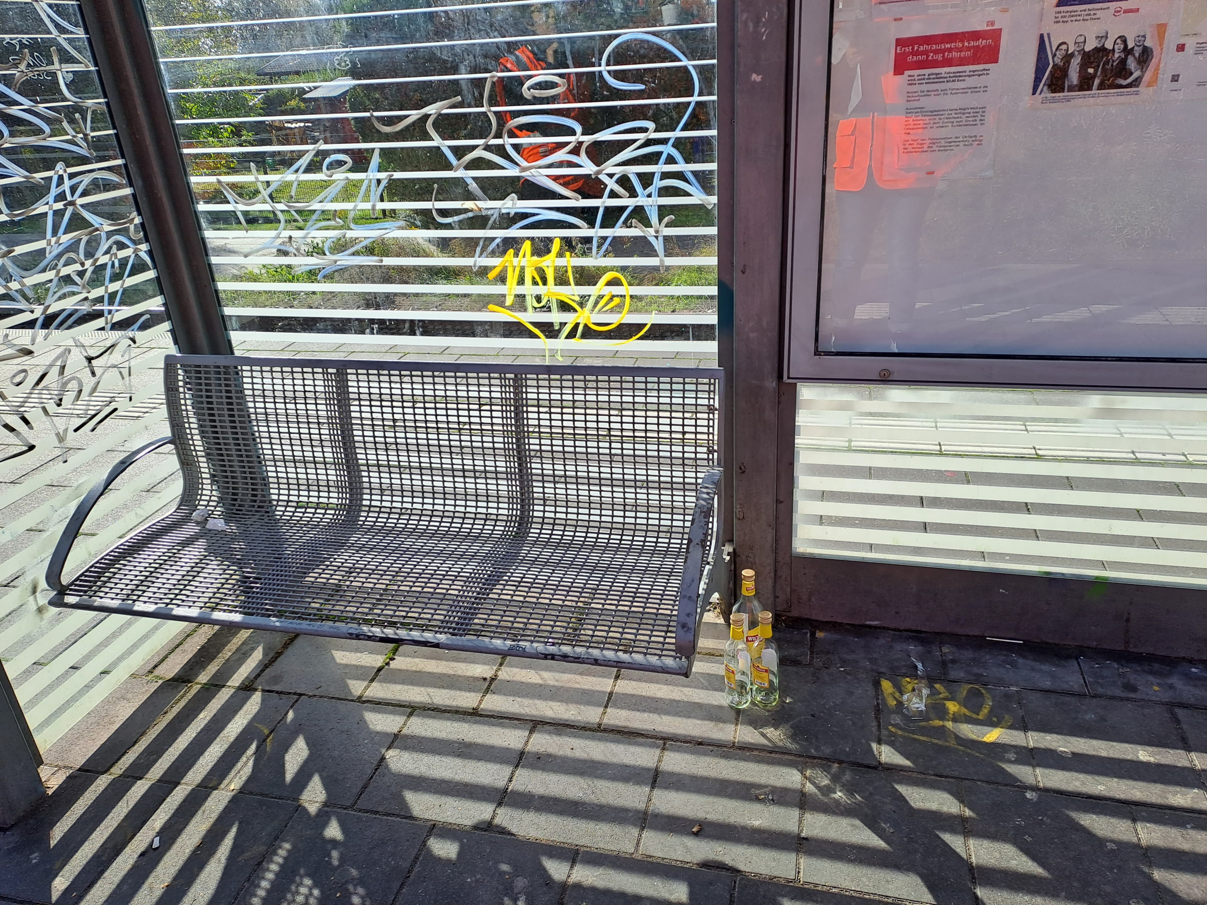 Eine graue Sitzbank in einem Wetterschutzhaus aus Glas und Stahl.