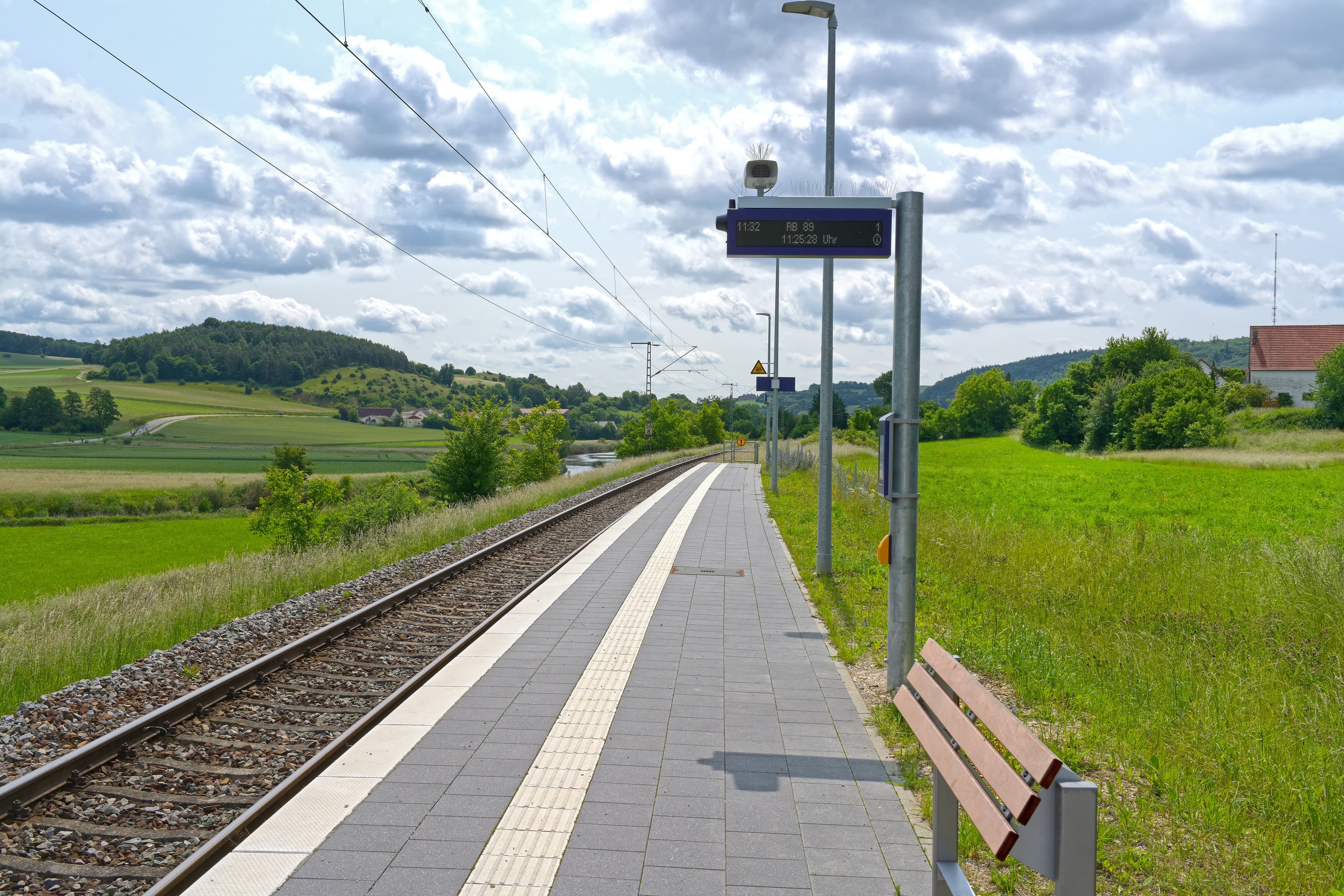 In der Mitte der Bahnsteig und links die Gleise. Rechts im Bild ein modernes Lehnmöbel aus Holz und Metall. Darüber der neue dynamische Schriftanzeiger.