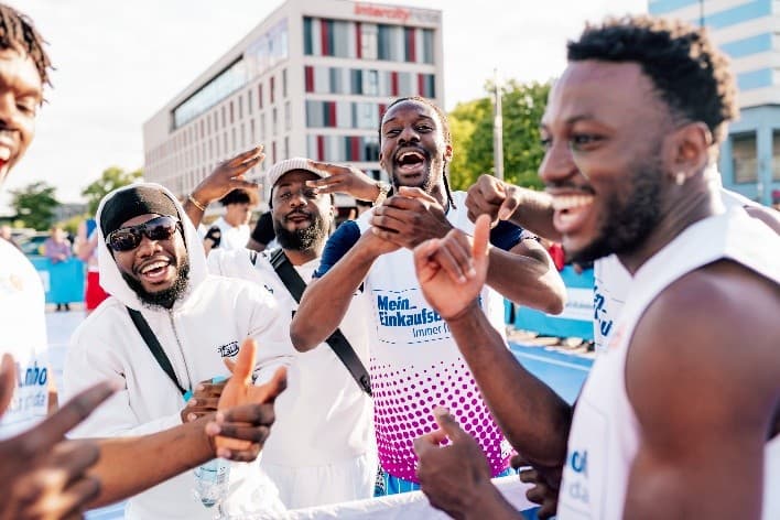 Mehrere lachende Männer in Sportkleidung stehen eng beieinander jubelnd auf einem Outdoor-Basketballcourt, während im Hintergrund moderne Gebäude zu sehen sind.