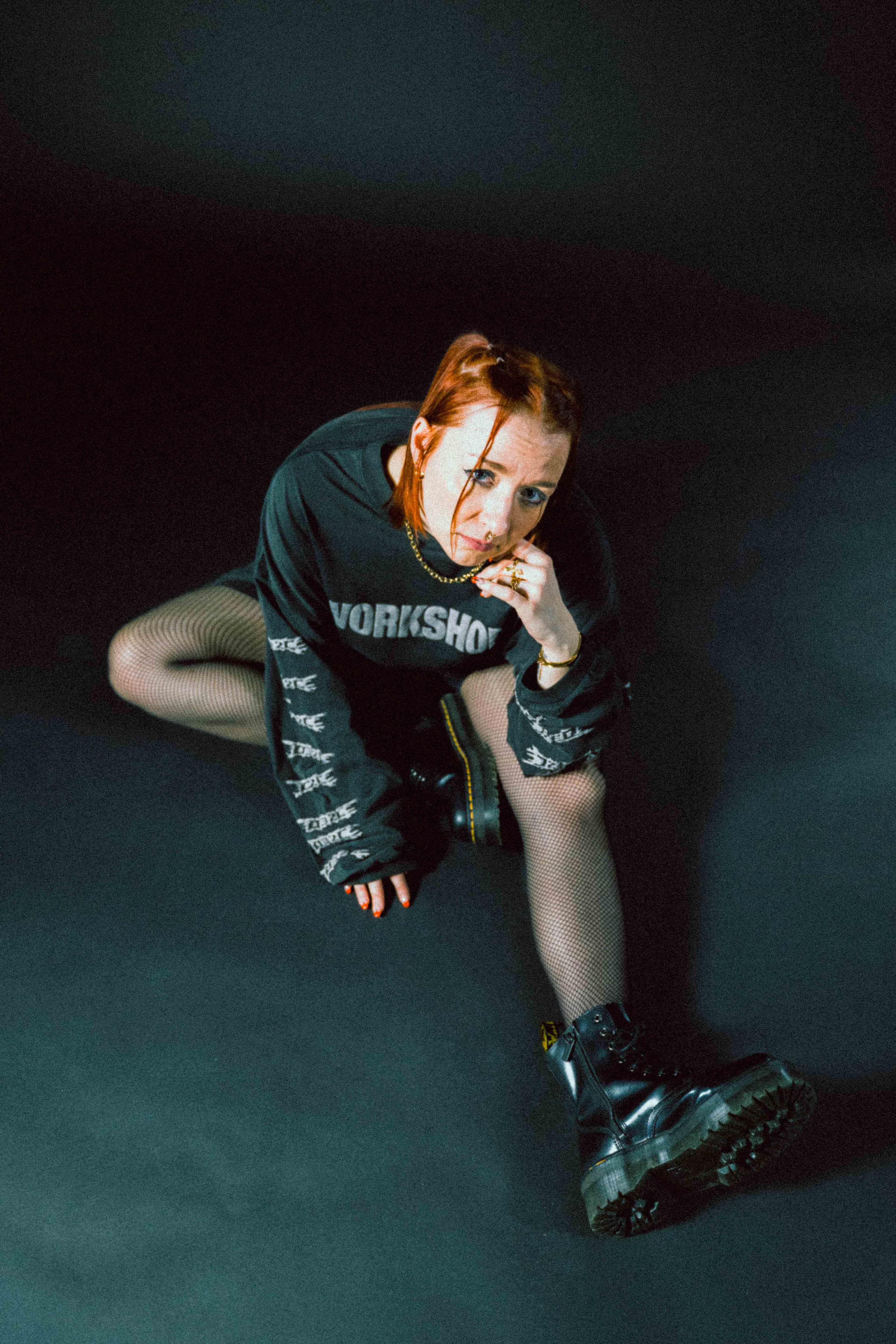 Jenoveva in schwarzem Kapuzenpullover mit weißem Schriftzug und Netzstrumpfhose sitzt mit einem ausgestreckten Bein auf dem Boden und trägt schwarze Stiefel