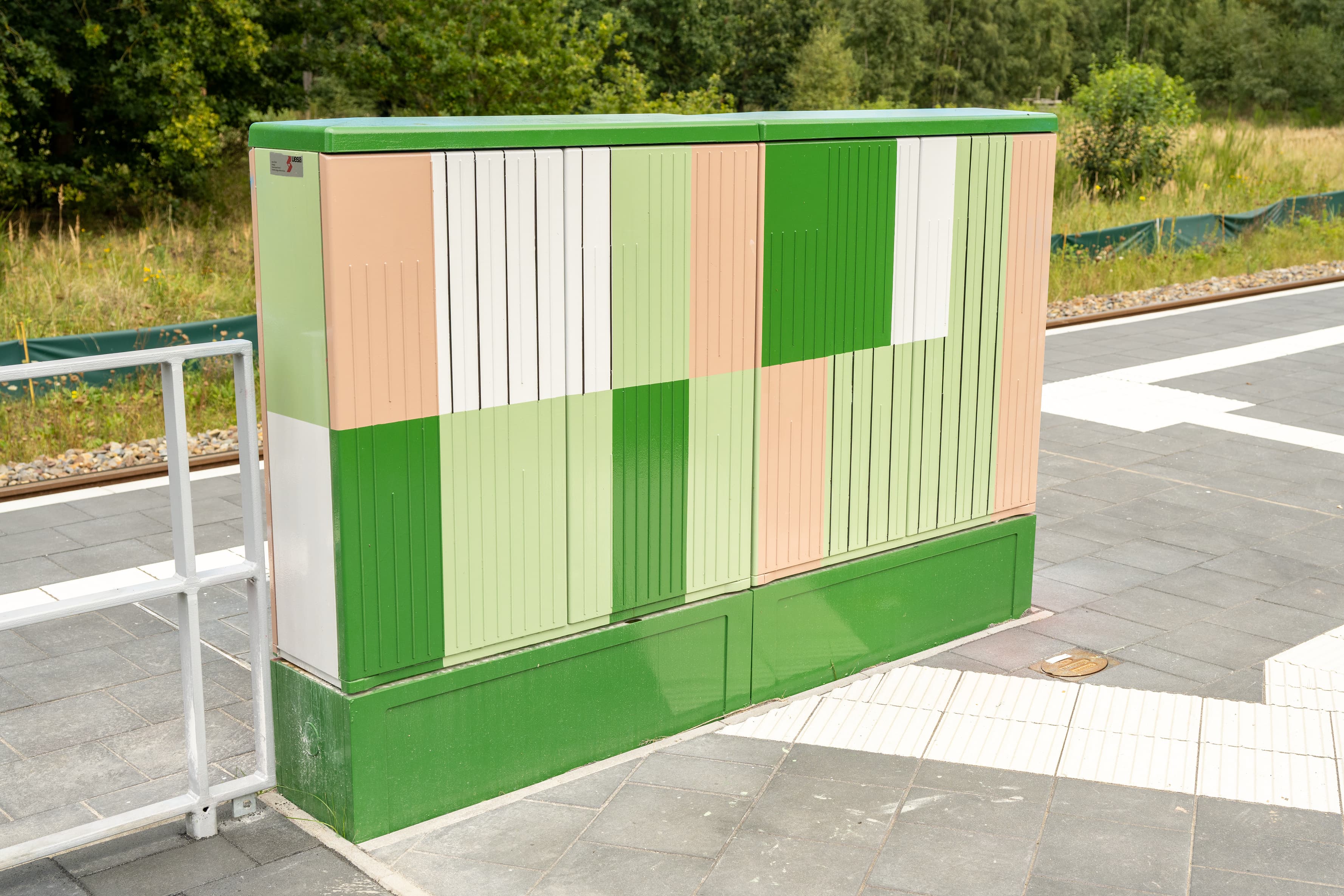 Großaufnahme eines Stromkastens auf dem Bahnsteig. Statt dem üblichen Lichtgrau ist dieses Exemplar mit einem geometrischen Muster in mehreren Grün- und Beigetönen versehen.