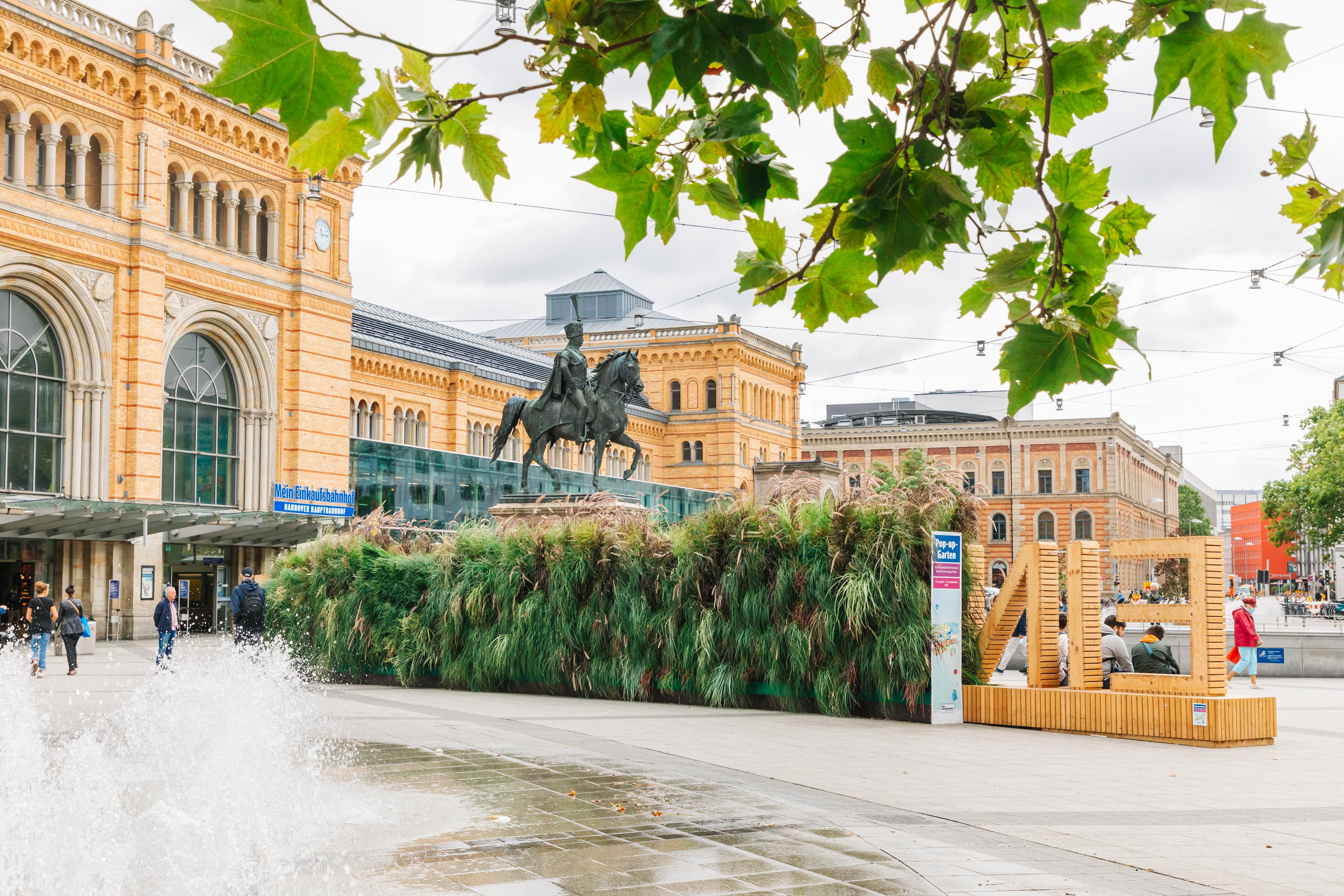 Blick auf die bepflanzte Rückwand des hölzernen Einkaufsbahnhof-Schriftzugs. Im Hintergrund ist das historische Bahnhofsgebäude des Hannover Hbf und das Pferde-Denkmal von Ernst-August zu sehen. Im Vordergrund sieht man den Brunnen auf dem Bahnhofsvorplatz.