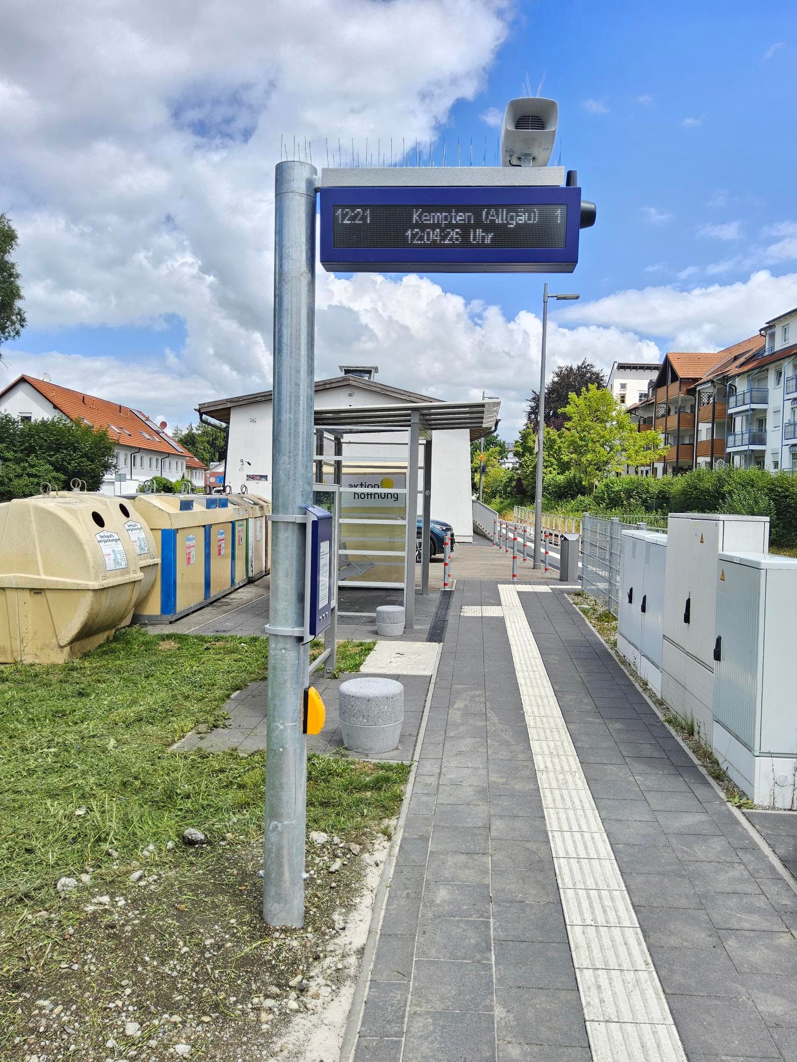 Links im Bild ein Mast mit einem Taster und darüber einem kleinen Bildschirm. Oben am Mast ein Bildschirm mit zwei Zeilen. Rechts darunter der Bahnsteig mit taktilem Leitsystem.