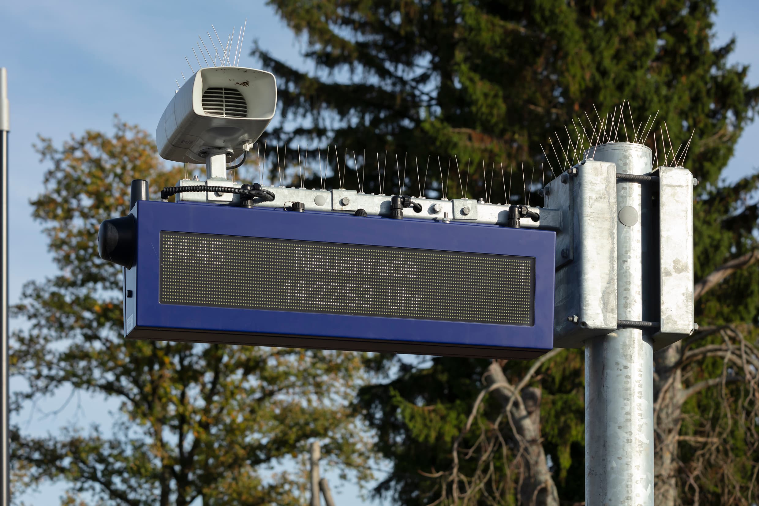 Dynamischer Schriftenanzeiger mit blauem Rand. Oben drauf ein Lautsprecher. Im Hintergrund Bäume.