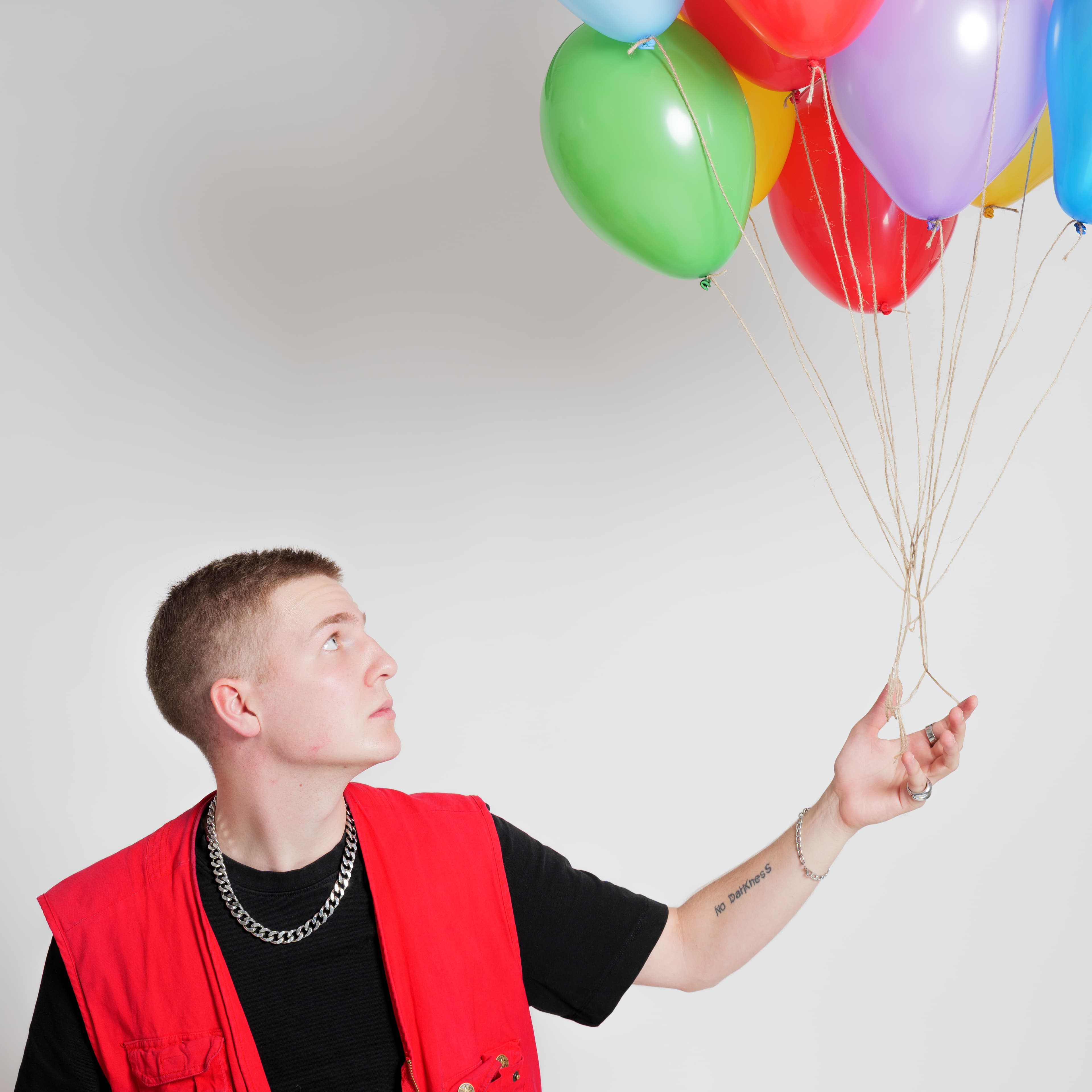Mosbach aus Wain mit kurzem Haar trägt rotes ärmelloses Oberteil über schwarzem T-Shirt und hält eine Gruppe bunter Luftballons in der linken Hand
